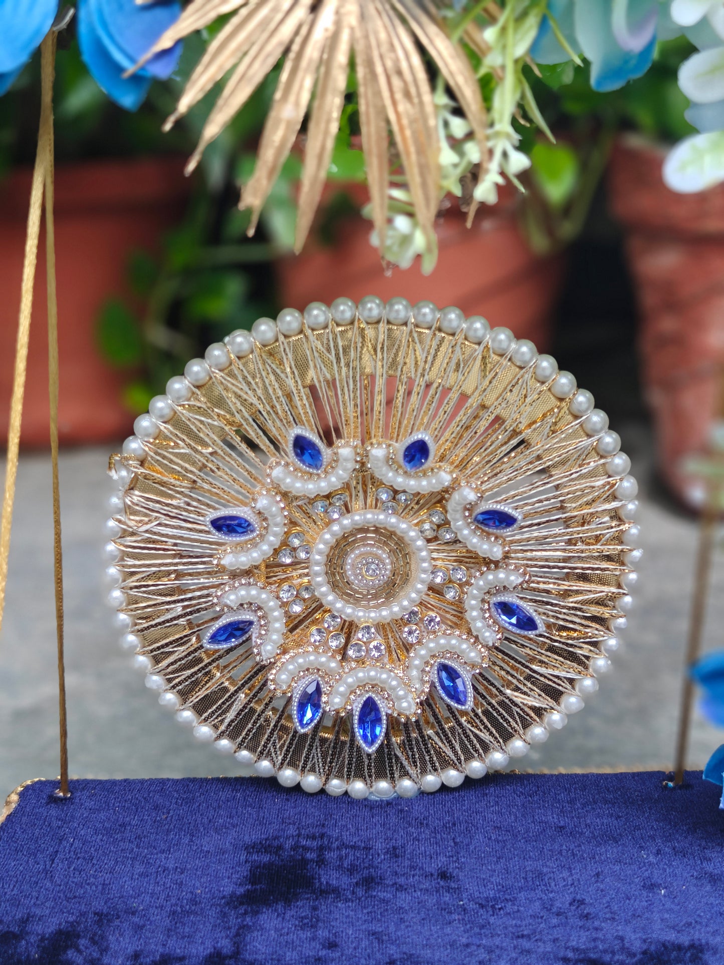 A round gold and white pearl embellished clutch bag with blue gemstone accents, displayed on a dark blue velvet surface with a blurred background of potted plants.