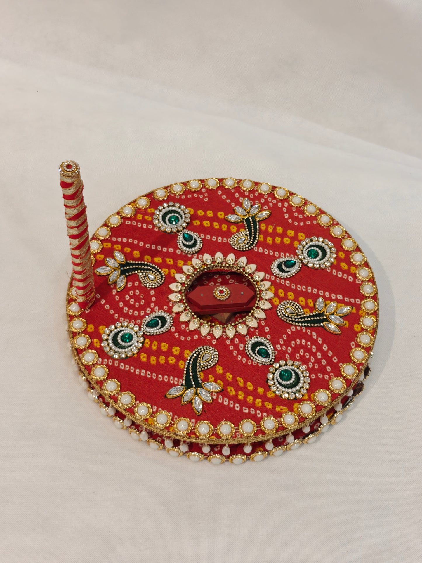 Side view of handcrafted red Bandhani decorative Baan chakki showing white pearl cabochon edge border, hanging pearl teardrop fringe underside detail and red white spiral ribbon wrapped handle with kundan finial on 12 inch MDF grinding wheel for Indian wedding ceremony