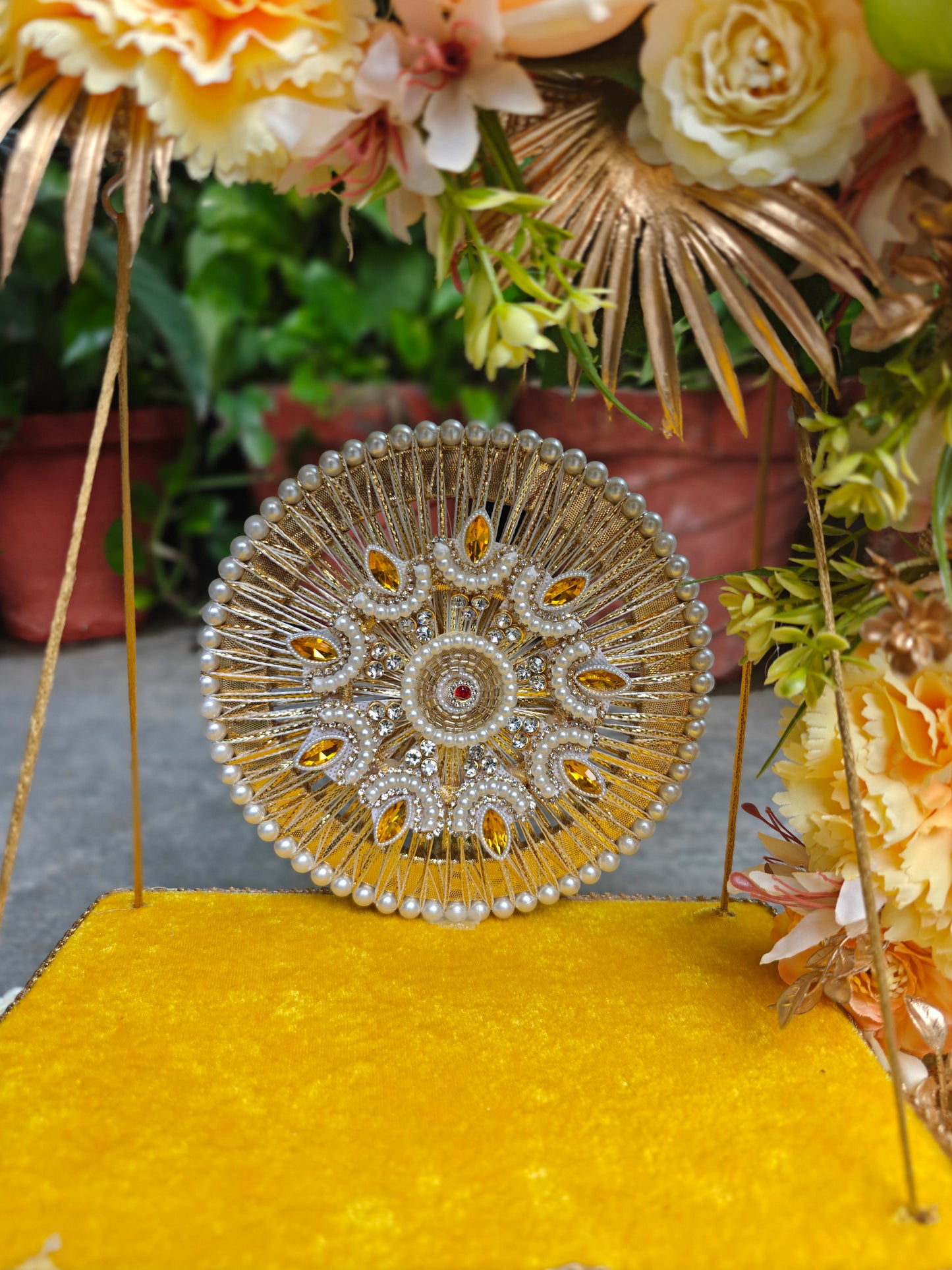A circular ornate gold and pearl decorative piece with intricate beadwork and small amber accents, displayed on a bright yellow surface with floral decor in the background.