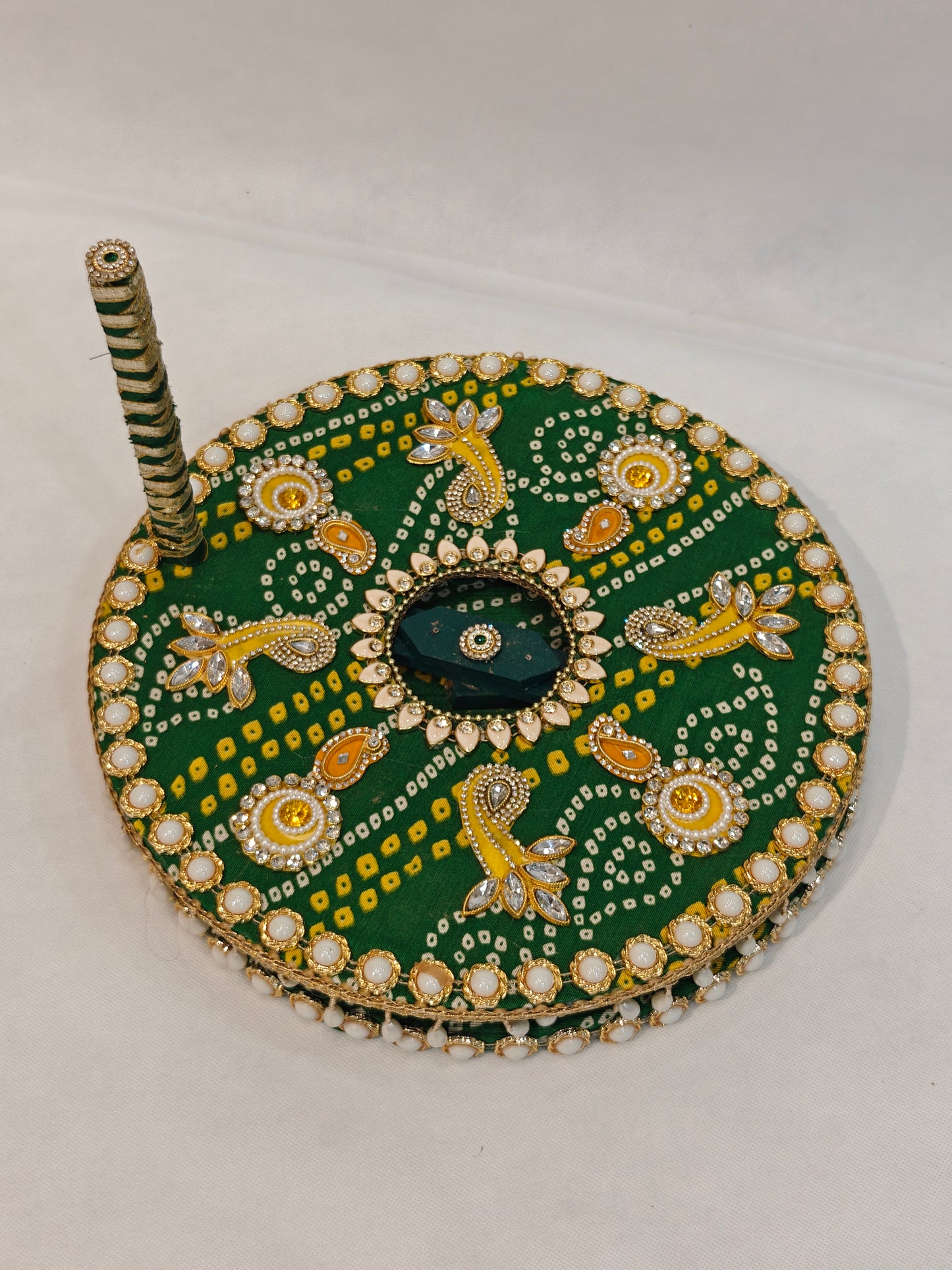 Side profile of handcrafted green Bandhani decorative Baan chakki showing white pearl cabochon edge border, hanging pearl teardrop fringe underside detail and green white spiral ribbon wrapped handle with kundan finial on 12 inch MDF grinding wheel for Indian pre-wedding ceremony