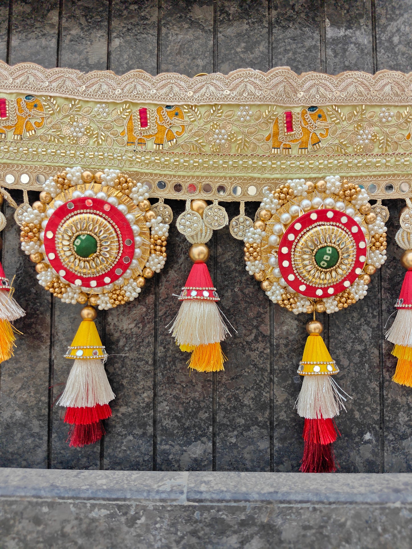 A close-up of an ornate embroidered textile decoration with a beige lace-trimmed band, gold elephant motifs, and two large circular embellishments in red, gold, white, and green, each hanging from tassels against a dark gray paneled background.