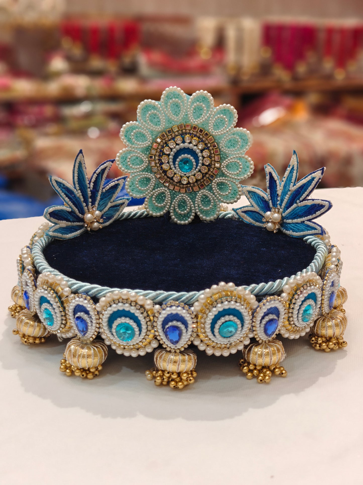 Handcrafted teal and blue lotus Singhaasan for Ladoo Gopal — 8 inch compact MDF deity throne with teal pearl floral medallion crown, twin royal blue lotus blooms, navy blue velvet seating, peacock-eye circle teardrop border and gold ghungroo tassel drops by The Wedding Station