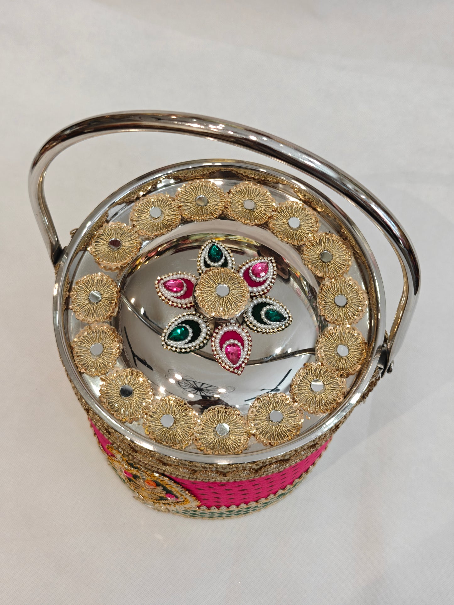 Top view of decorated stainless steel wedding bucket lid showing gold zardozi sunflower medallions with mirror insets and central handcrafted pink and green kundan teardrop floral motif with seed pearl outlines and arched steel handle