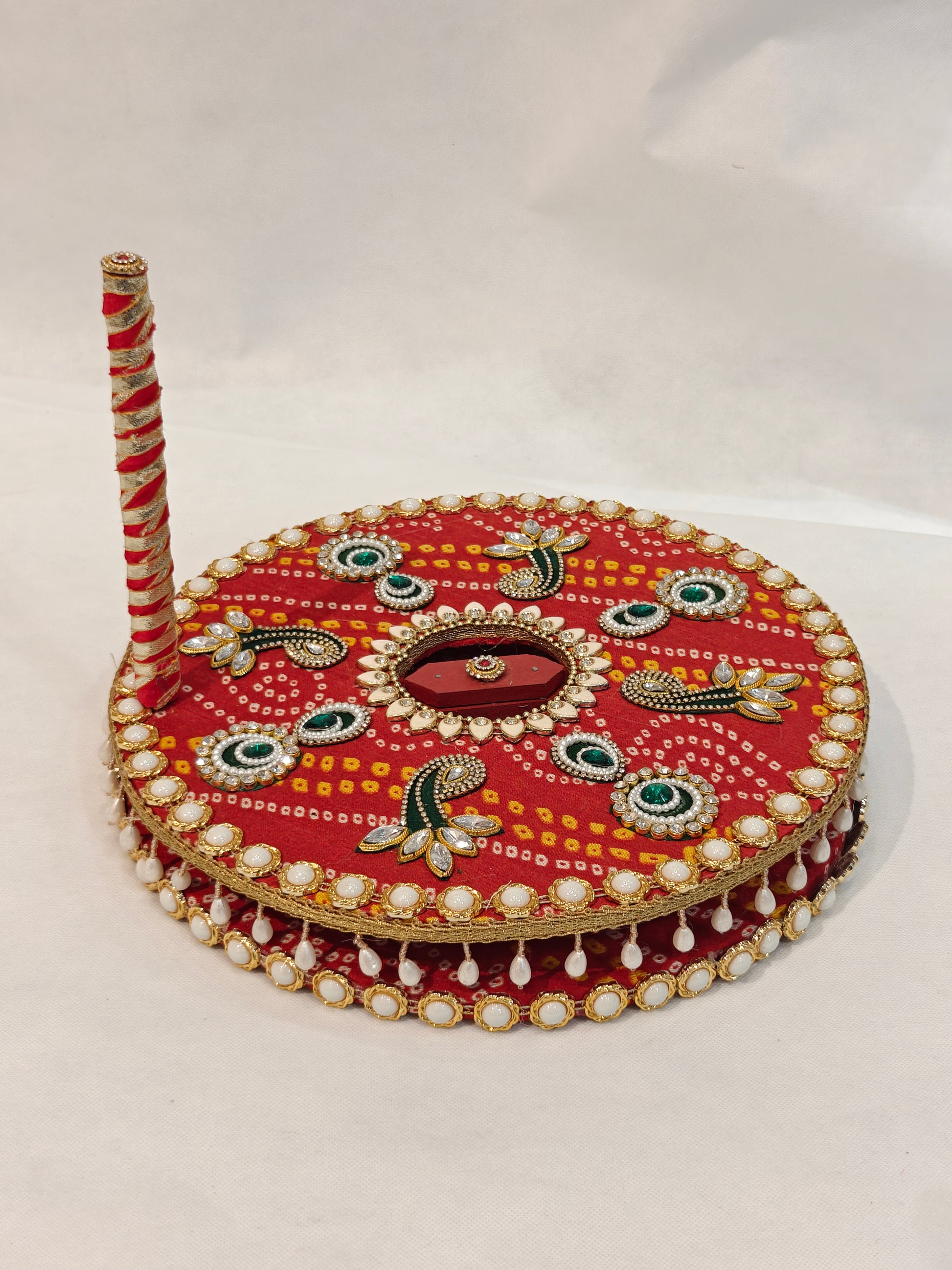 Decorative MDF chakki for Indian Baan pre-wedding ritual in red Bandhani fabric with green kundan eye motifs, black gold paisley, hexagonal mirror centre, white kundan lotus burst, pearl cabochon border, hanging pearl drop fringe and red white ribbon wrapped handle — 12 inch diameter