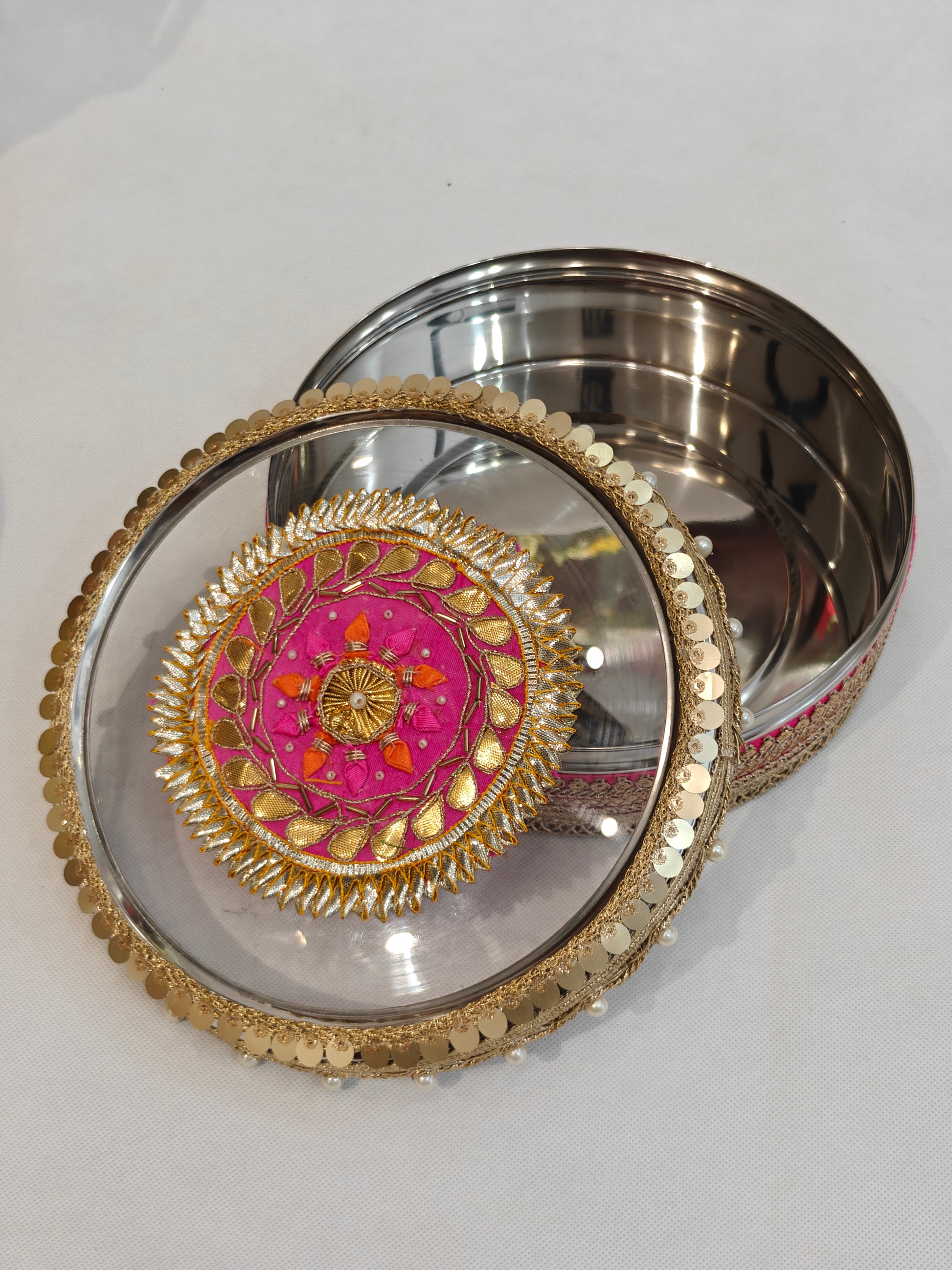 Open decorated stainless steel wedding storage box showing handcrafted zardozi embroidery medallion on hot pink base with gold gota patti, orange silk petals and pearl accents on transparent lid
