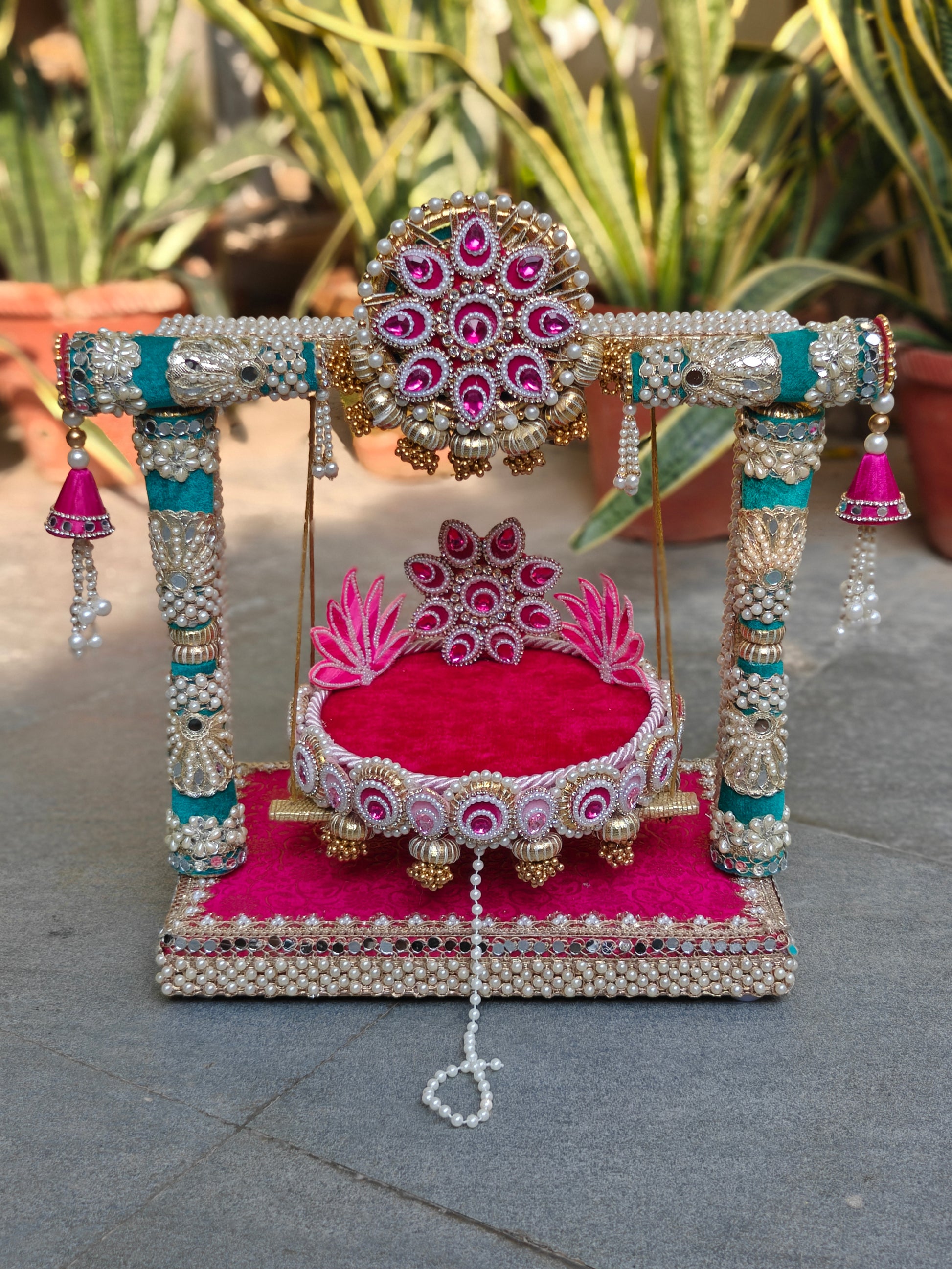 Royal Collection 2-in-1 handcrafted Ladoo Gopal jhula and singhasan in rani pink and teal velvet – decorative torana crown with fuchsia teardrop stones, teal pillar frame with mirror work and pearl clusters, detachable lotus patra singhasan with gold ghungroo fringe, MDF base with mirror border and pearl edging – The Wedding Station