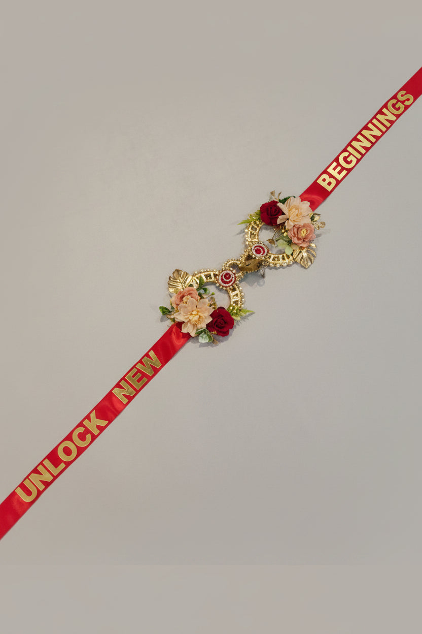 Handcrafted red satin inauguration ribbon printed with "Unlock New Beginnings" in gold text, with twin interlocked gold rings decorated with artificial red roses, blush peonies and crystal meenakari centrepiece — shop opening and housewarming welcome lock ribbon by The Wedding Station Meerut