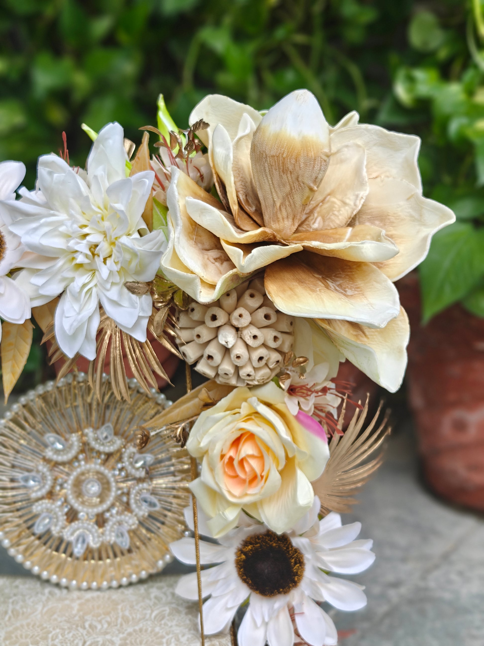 A decorative floral arrangement featuring large cream and tan magnolia-style flowers, white daisies, a peach rose, dried botanical elements, and a round beaded gold-toned medallion against a blurred green outdoor background.