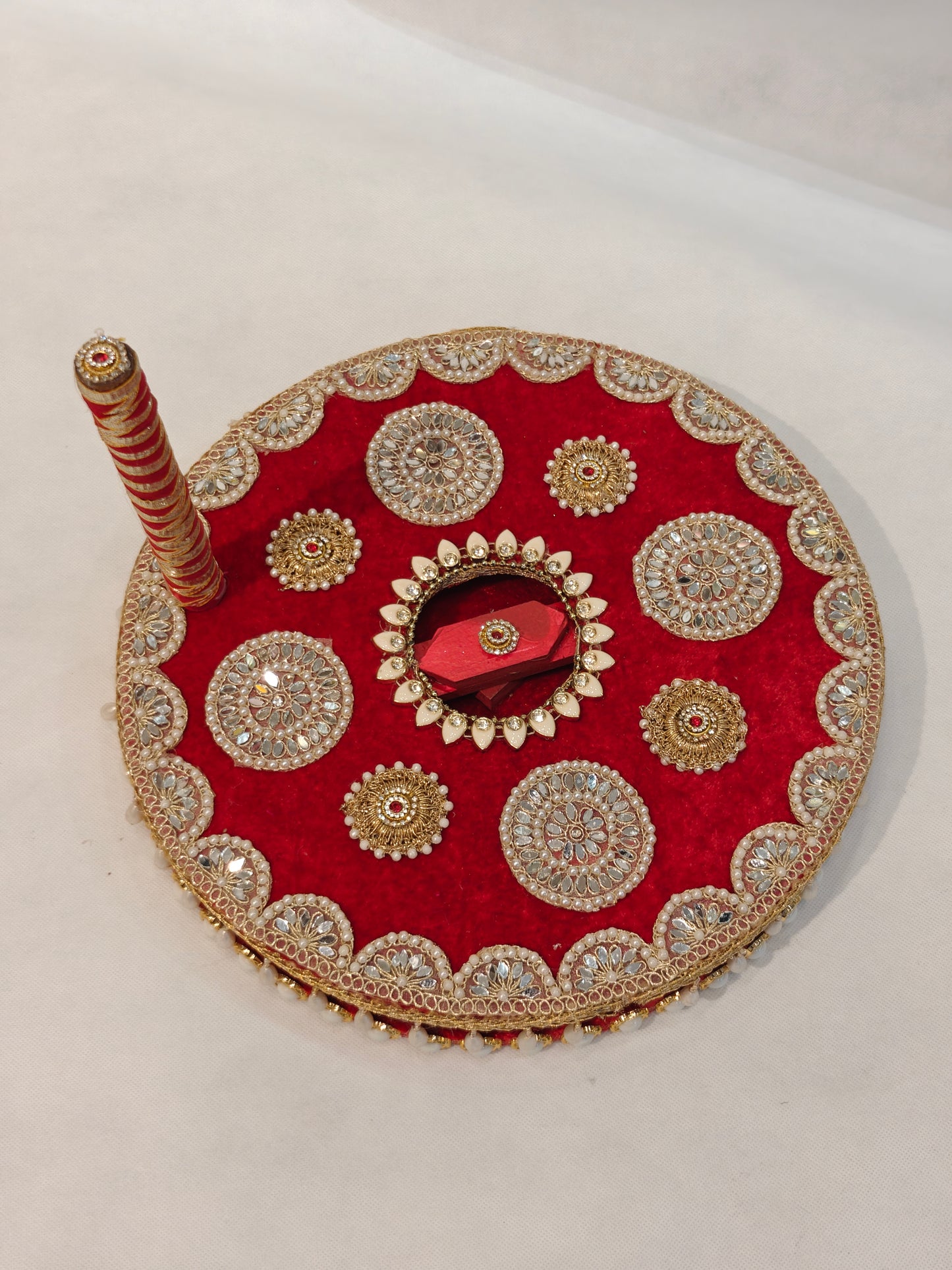 Decorative MDF chakki for Indian Baan pre-wedding ritual in deep red velvet with large circular mirror-work shisha medallions, gold zardozi sunburst motifs, red hexagonal mirror centre, white kundan lotus surround, gold scalloped edge lace and red gold ribbon wrapped handle — 12 inch diameter