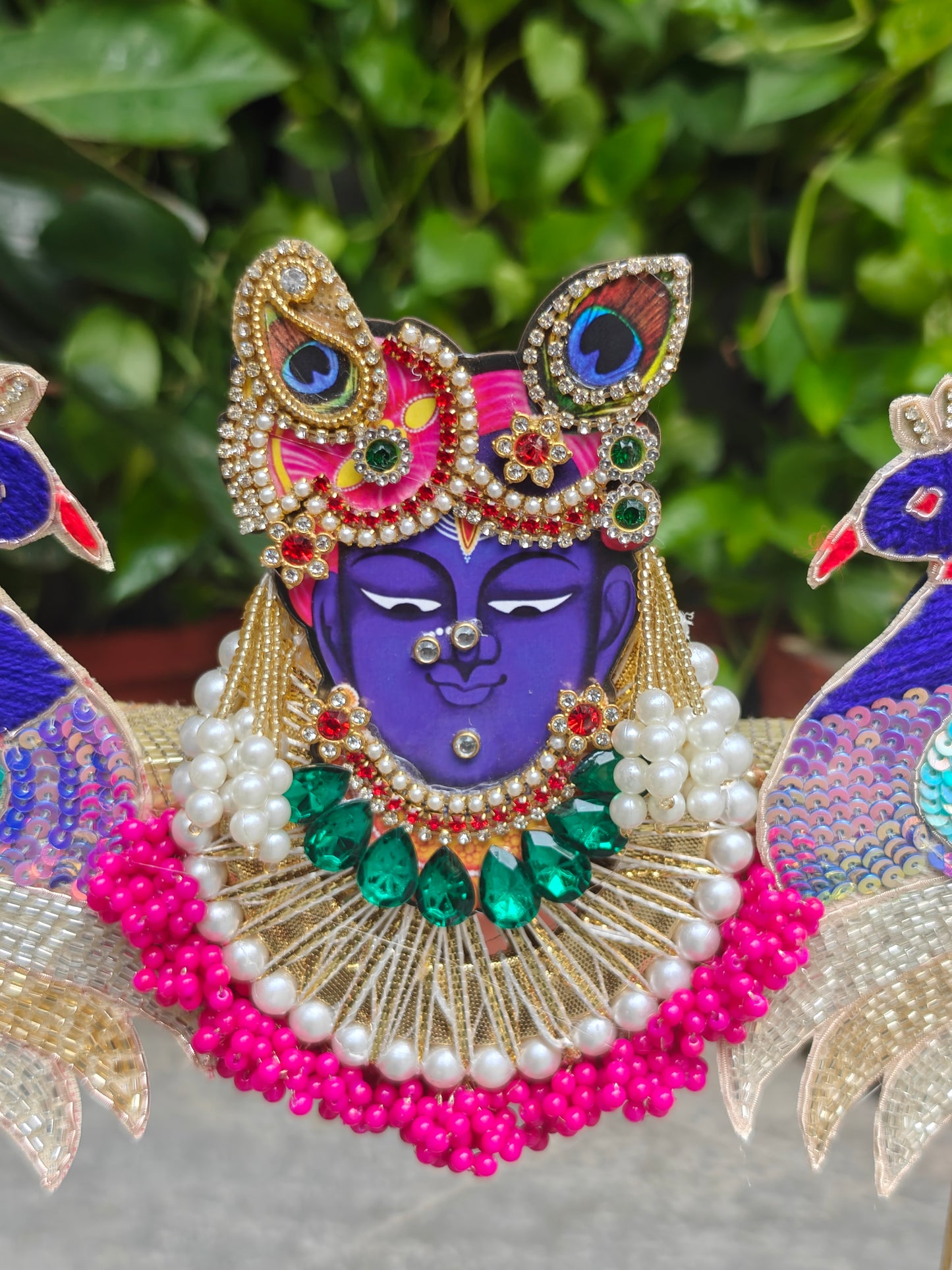 A close-up of an ornate decorative idol or wall hanging featuring a purple-skinned divine face with jeweled gold ornaments, pink and green embellishments, white pearl-like beads, and peacock motifs on both sides, set against a blurred green foliage background.