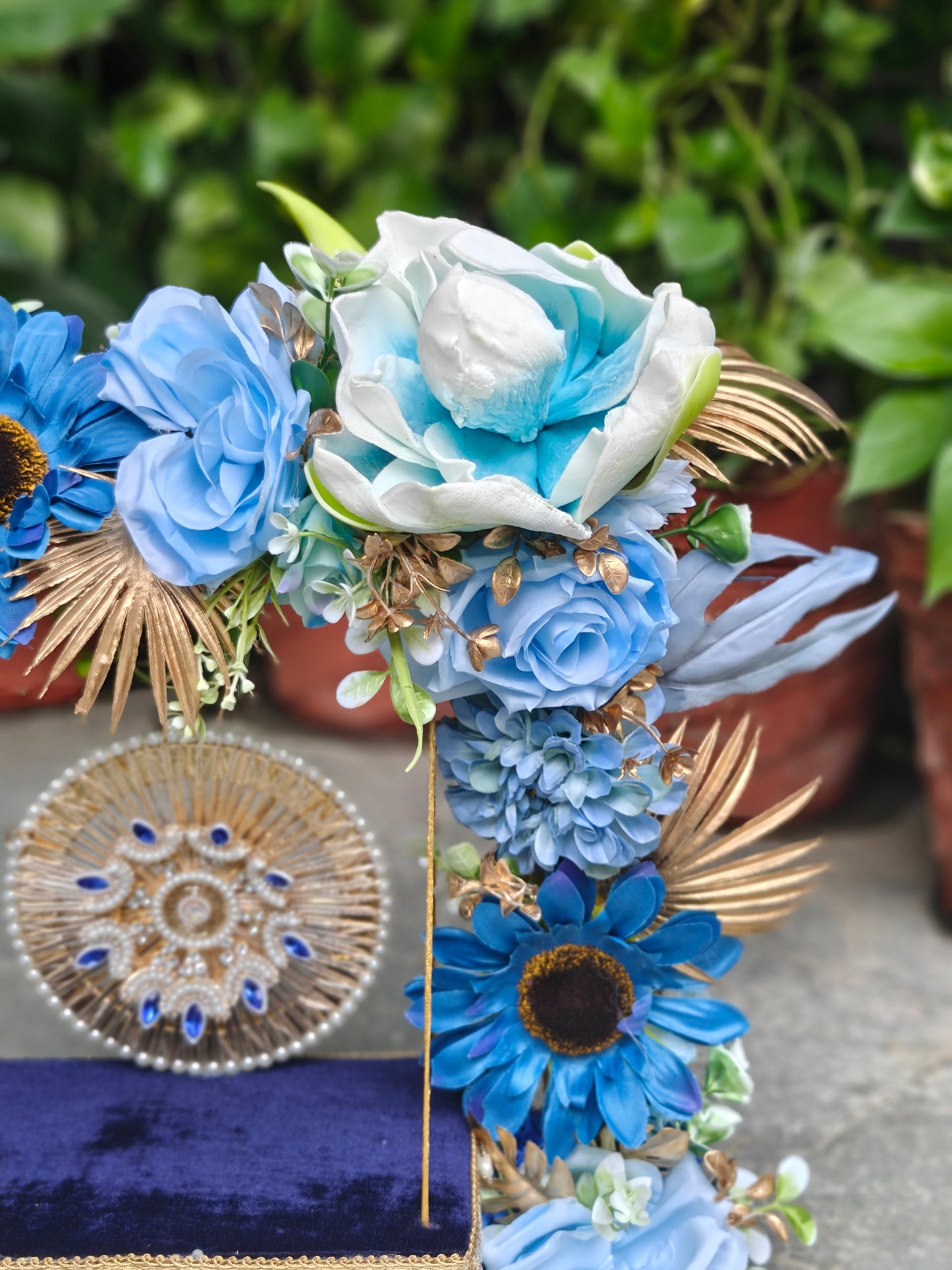 A decorative floral arrangement in shades of blue, white, and gold, styled with artificial flowers and leaves, displayed on a dark blue velvet surface with a round woven decorative piece beside it. The background shows blurred green foliage and terracotta pots.