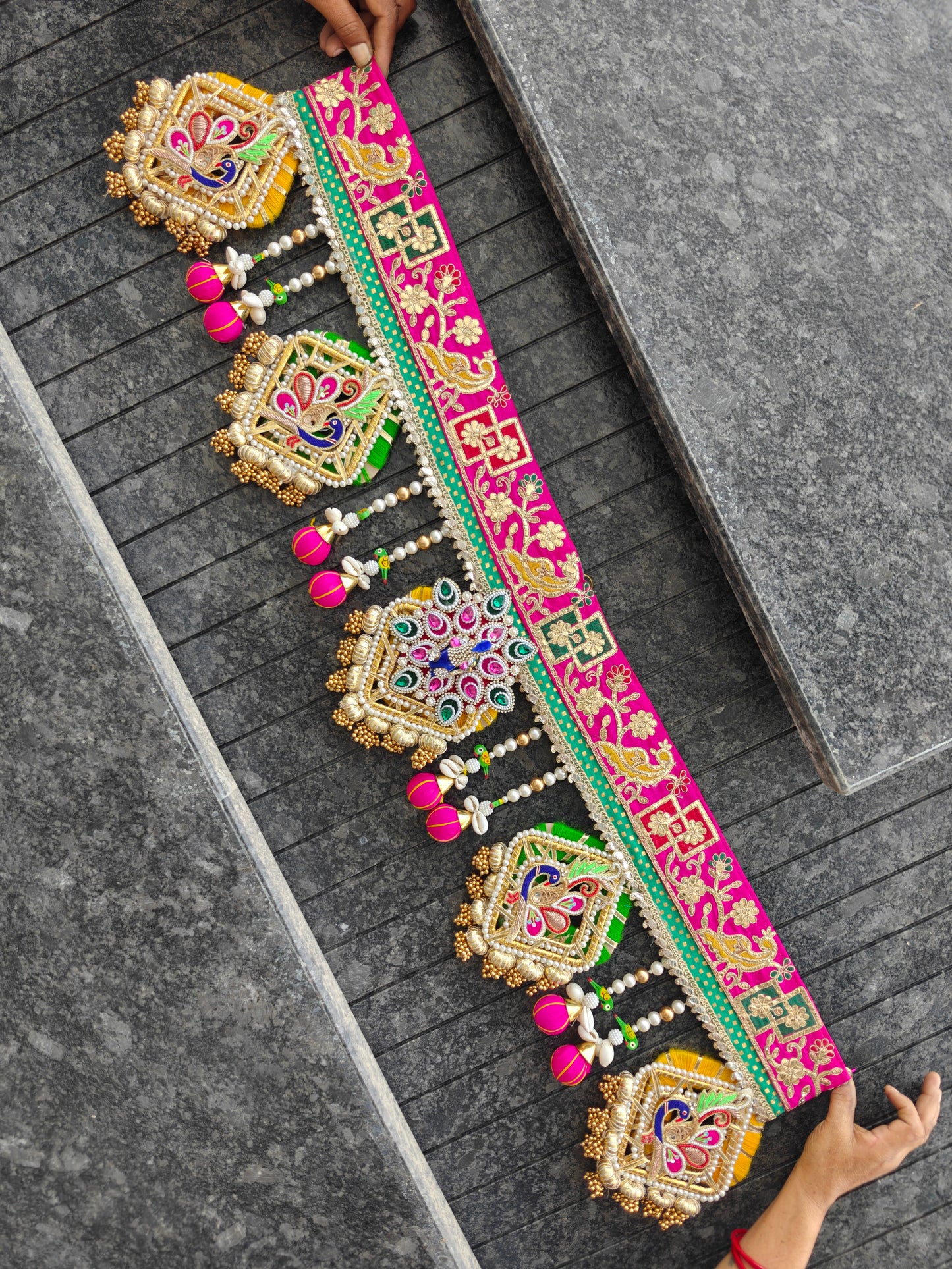 A long, ornate embroidered decorative belt or sash is spread across dark gray stone steps, held at both ends by hands. The belt features bright pink borders, intricate gold, green, and blue embroidery, and dangling pink tassel ornaments.