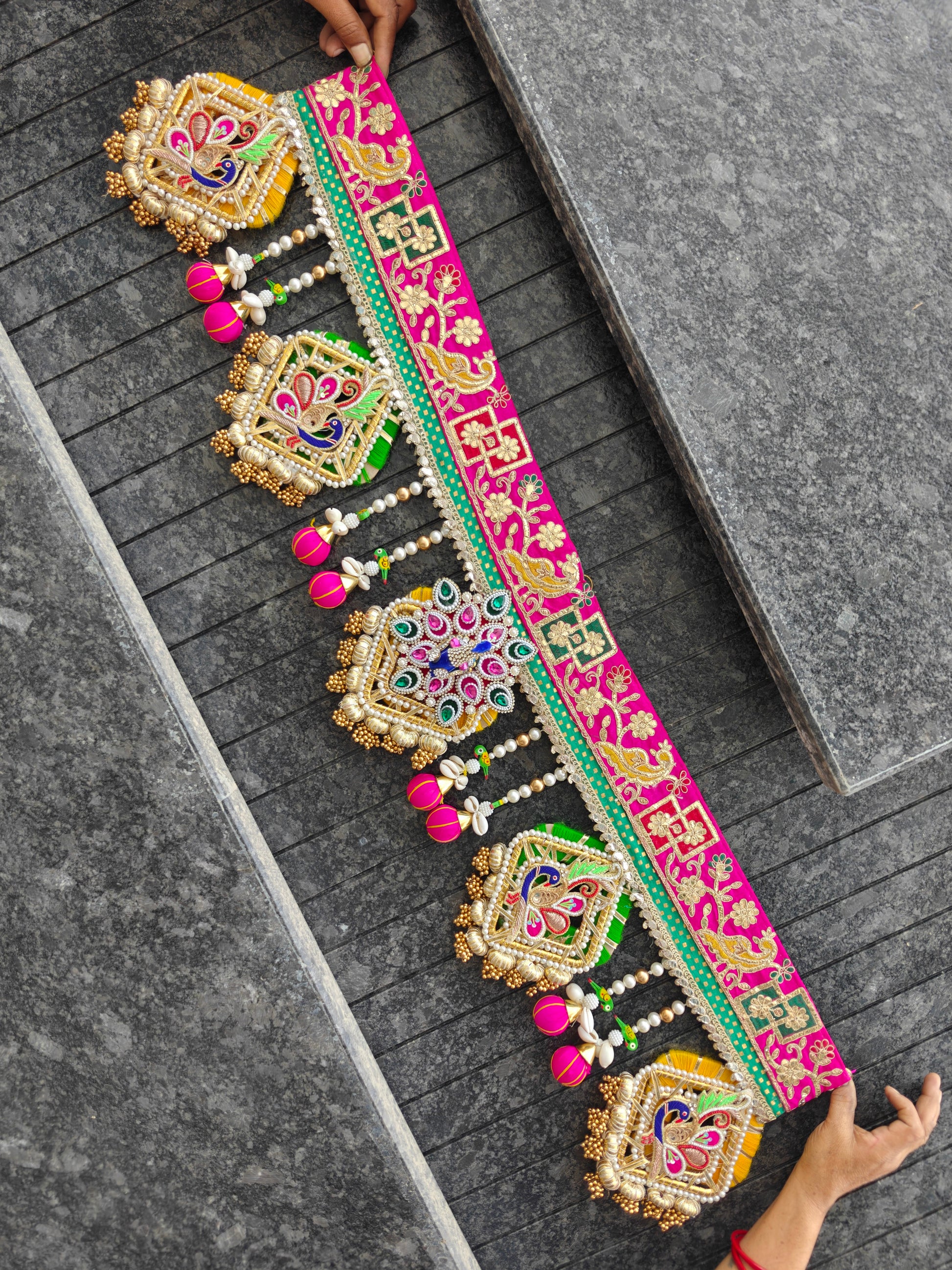 A long, ornate embroidered decorative belt or sash is spread across dark gray stone steps, held at both ends by hands. The belt features bright pink borders, intricate gold, green, and blue embroidery, and dangling pink tassel ornaments.