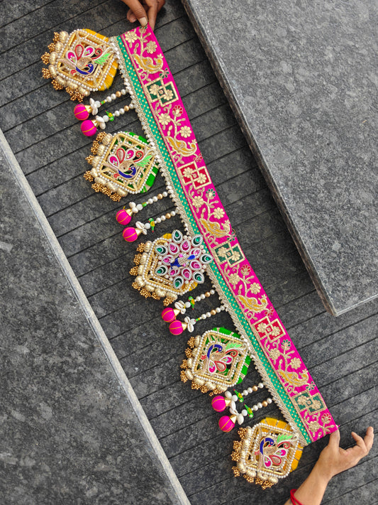A long, ornate embroidered decorative belt or sash is spread across dark gray stone steps, held at both ends by hands. The belt features bright pink borders, intricate gold, green, and blue embroidery, and dangling pink tassel ornaments.