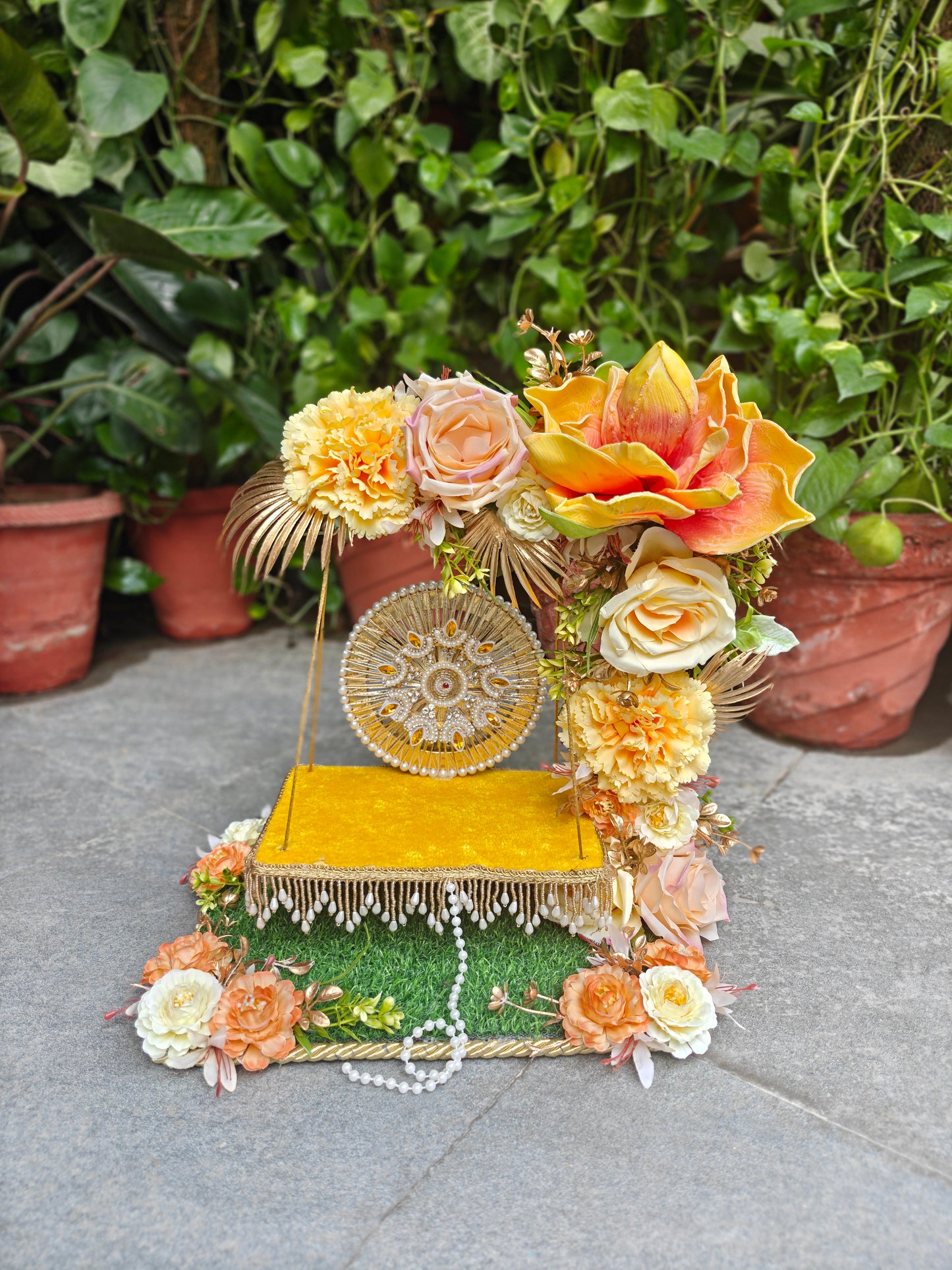 A decorative miniature golden stage set with a yellow velvet-like platform, floral arrangements in peach, cream, and orange, a round gold medallion, faux grass, and white bead garland, photographed outdoors on a stone surface with potted greenery in the background.