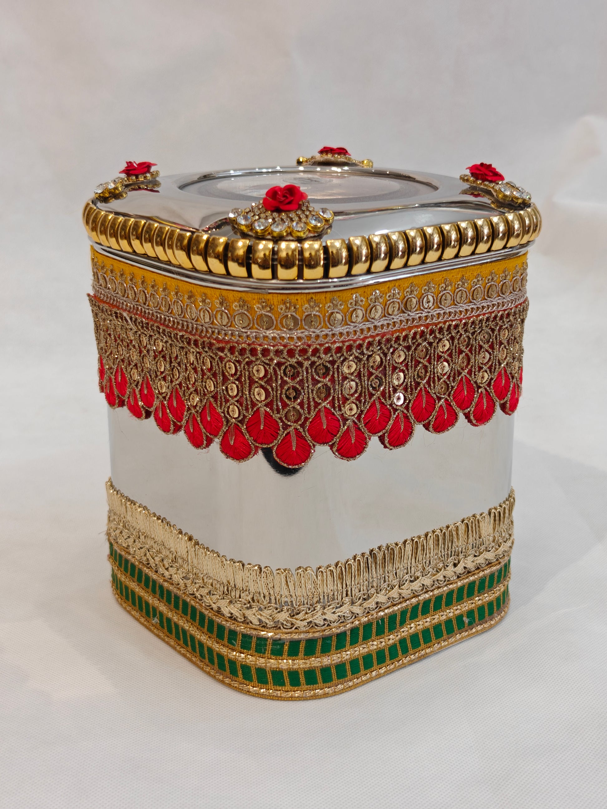 Decorated square stainless steel storage box for Indian wedding with red and gold scalloped zari lace, gold fringe band, green border, kundan rose corner accents and transparent acrylic lid window — 5.5x5.5 inch base, 7 inch height