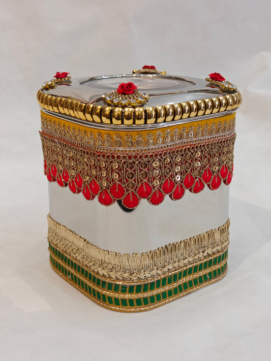 Decorated square stainless steel storage box for Indian wedding with red and gold scalloped zari lace, gold fringe band, green border, kundan rose corner accents and transparent acrylic lid window — 5.5x5.5 inch base, 7 inch height