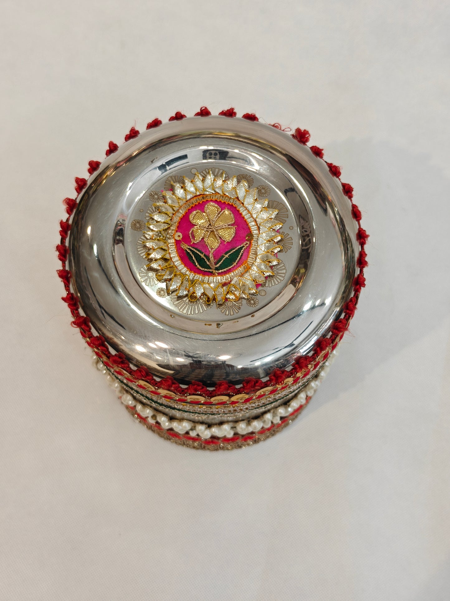Top view of decorated stainless steel round wedding box lid featuring handcrafted gota patti floral medallion on hot pink base with gold motif and green silk leaves, encircled by silver and gold petals with red tassel edge trim