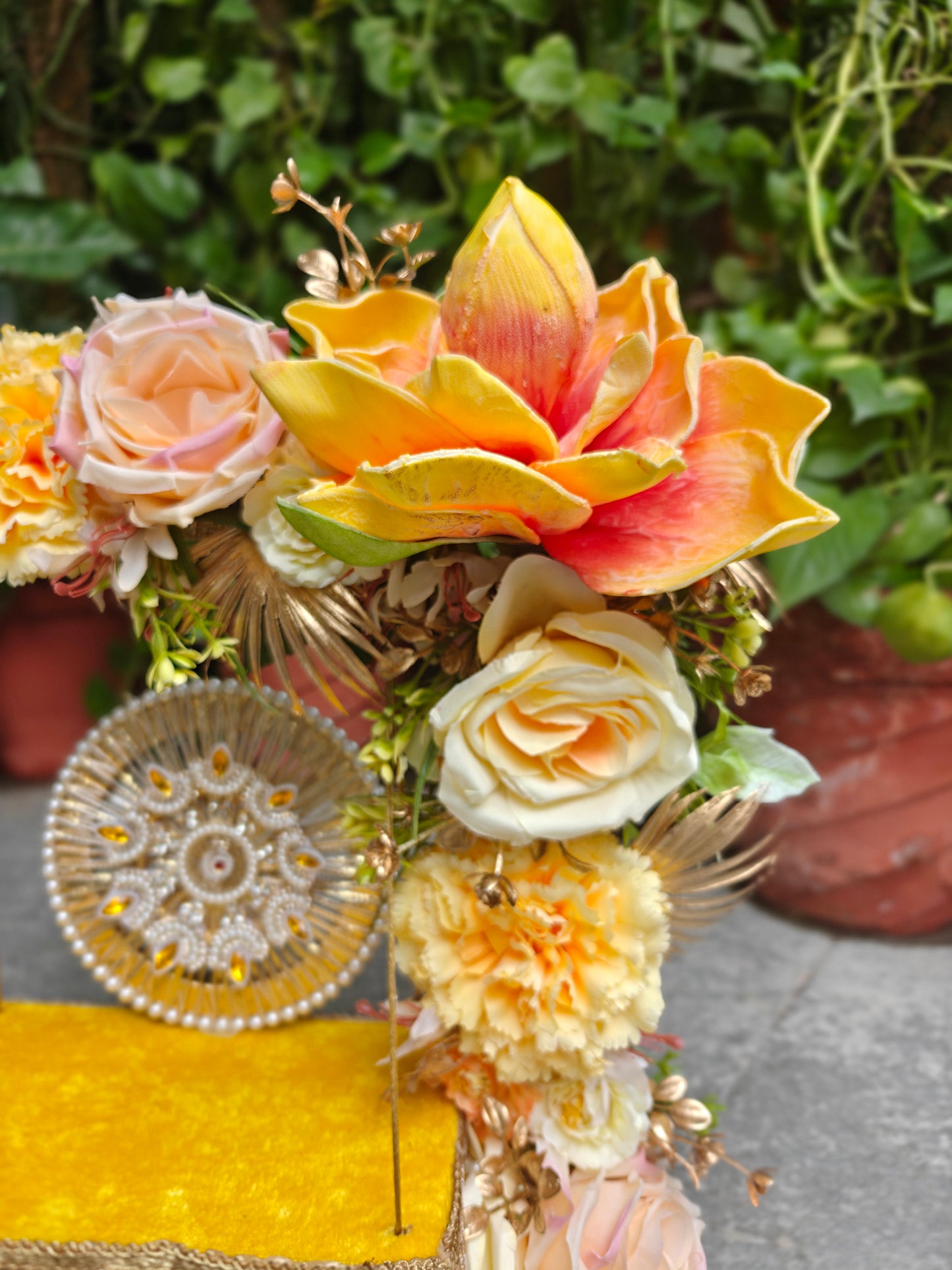 A decorative floral arrangement featuring a large yellow and orange lotus flower, peach roses, cream roses, and marigold-like blooms, styled with gold-toned accents and a round patterned plate on a bright yellow surface, with a blurred green garden background.
