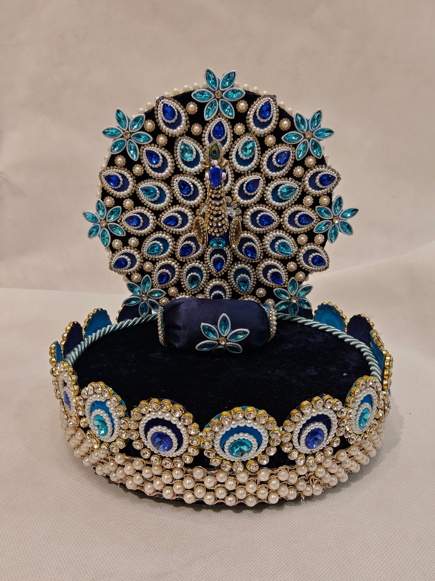 Front view of peacock Singhaasan for Ladoo Gopal — 10 inch handcrafted MDF deity throne with full-spread peacock backdrop, teardrop gem feather detailing, rhinestone border and pearl base by The Wedding Station