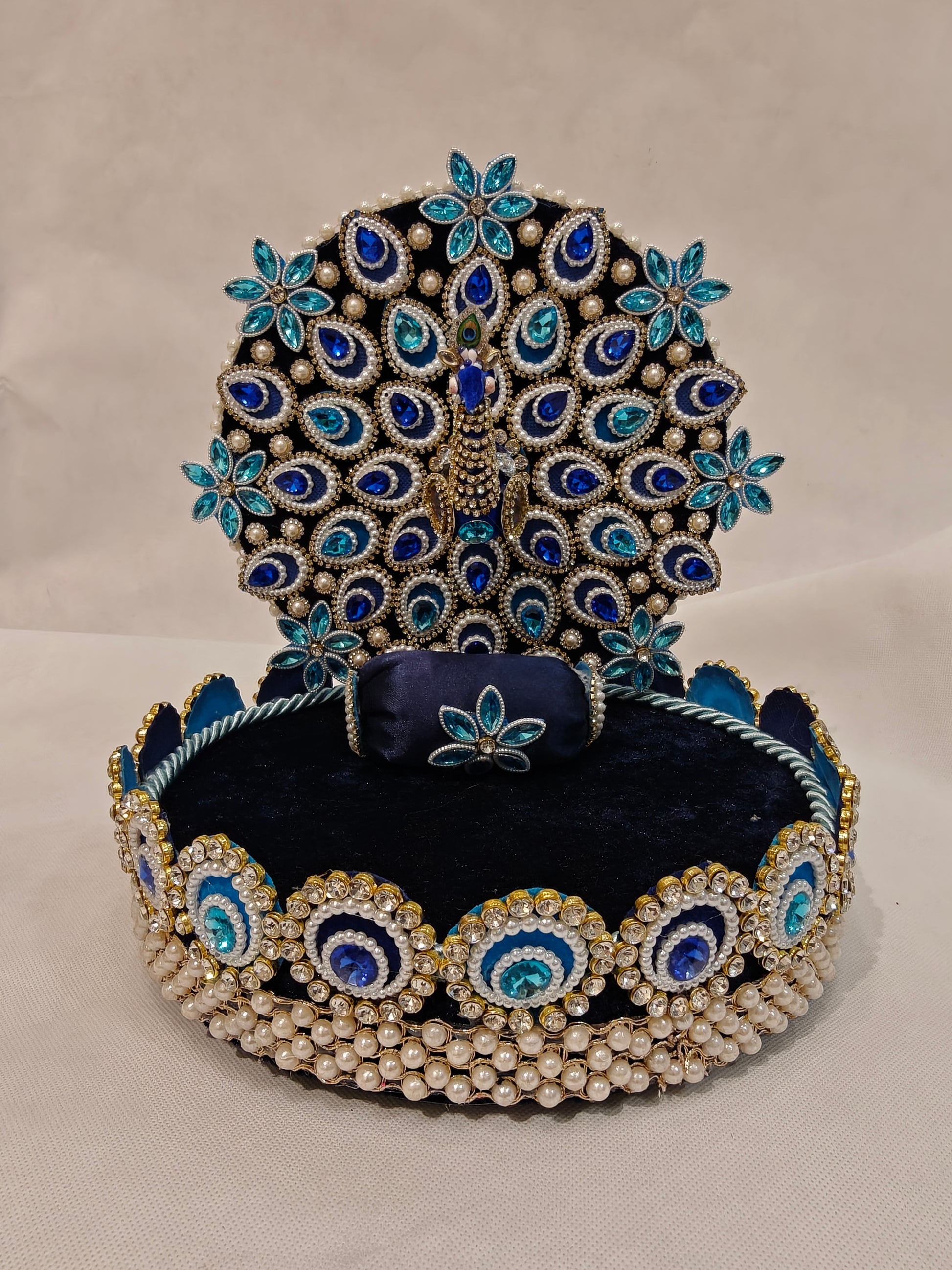 Front view of peacock Singhaasan for Ladoo Gopal — 10 inch handcrafted MDF deity throne with full-spread peacock backdrop, teardrop gem feather detailing, rhinestone border and pearl base by The Wedding Station