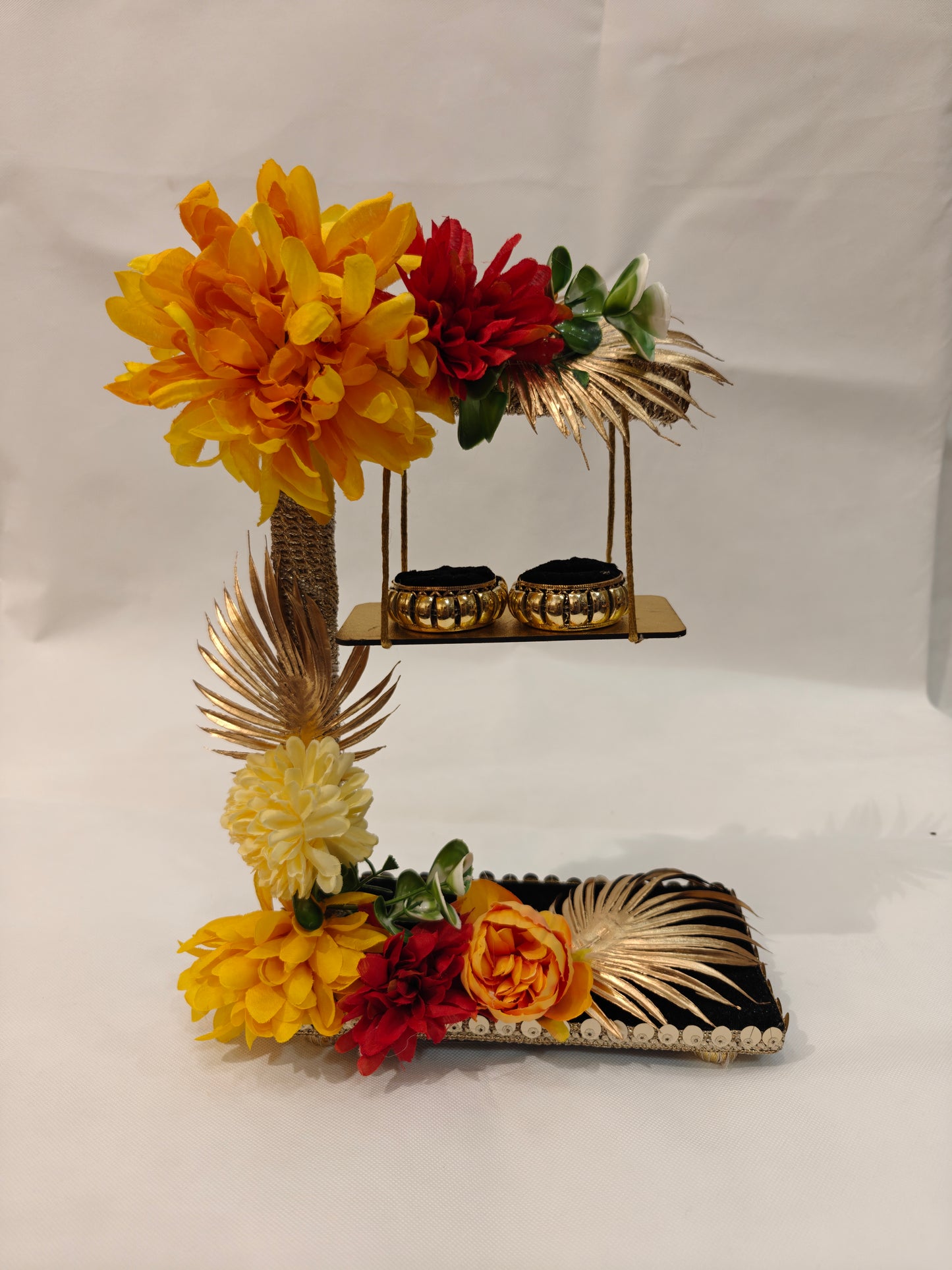2-tier handcrafted wooden ring ceremony stand with yellow, red, and orange silk flowers, gold palm leaves, jute-wrapped column, swing shelf, and two gold ring holders — by The Wedding Station