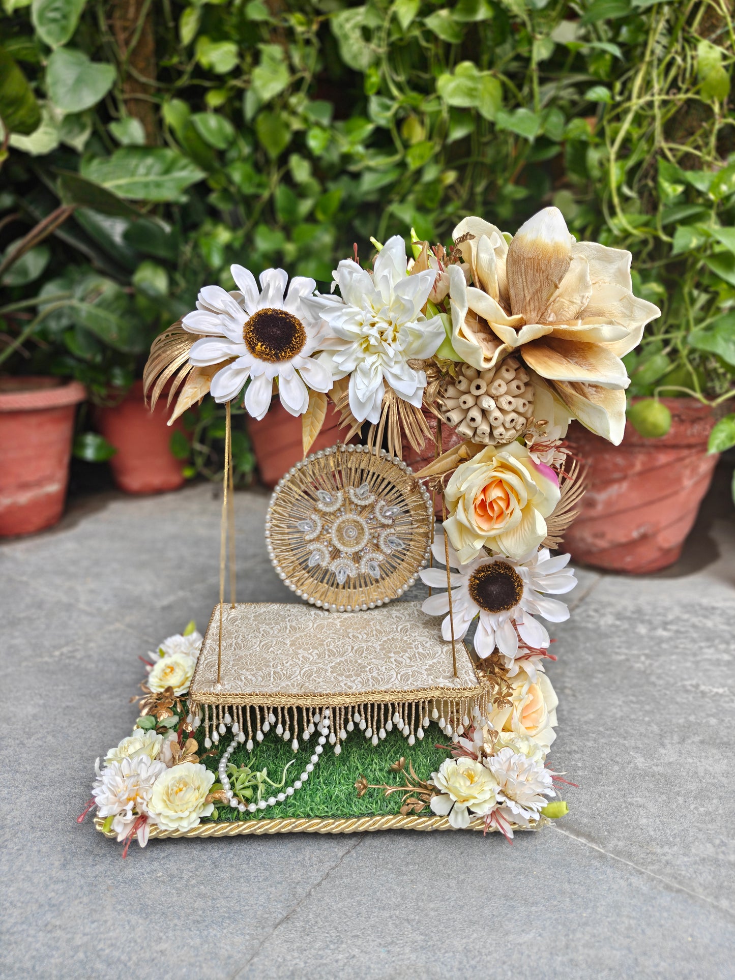 A decorative floral display stand with faux flowers, a small woven basket, faux pearls, and a lace-trimmed platform, photographed outdoors on a stone surface with potted plants in the background.