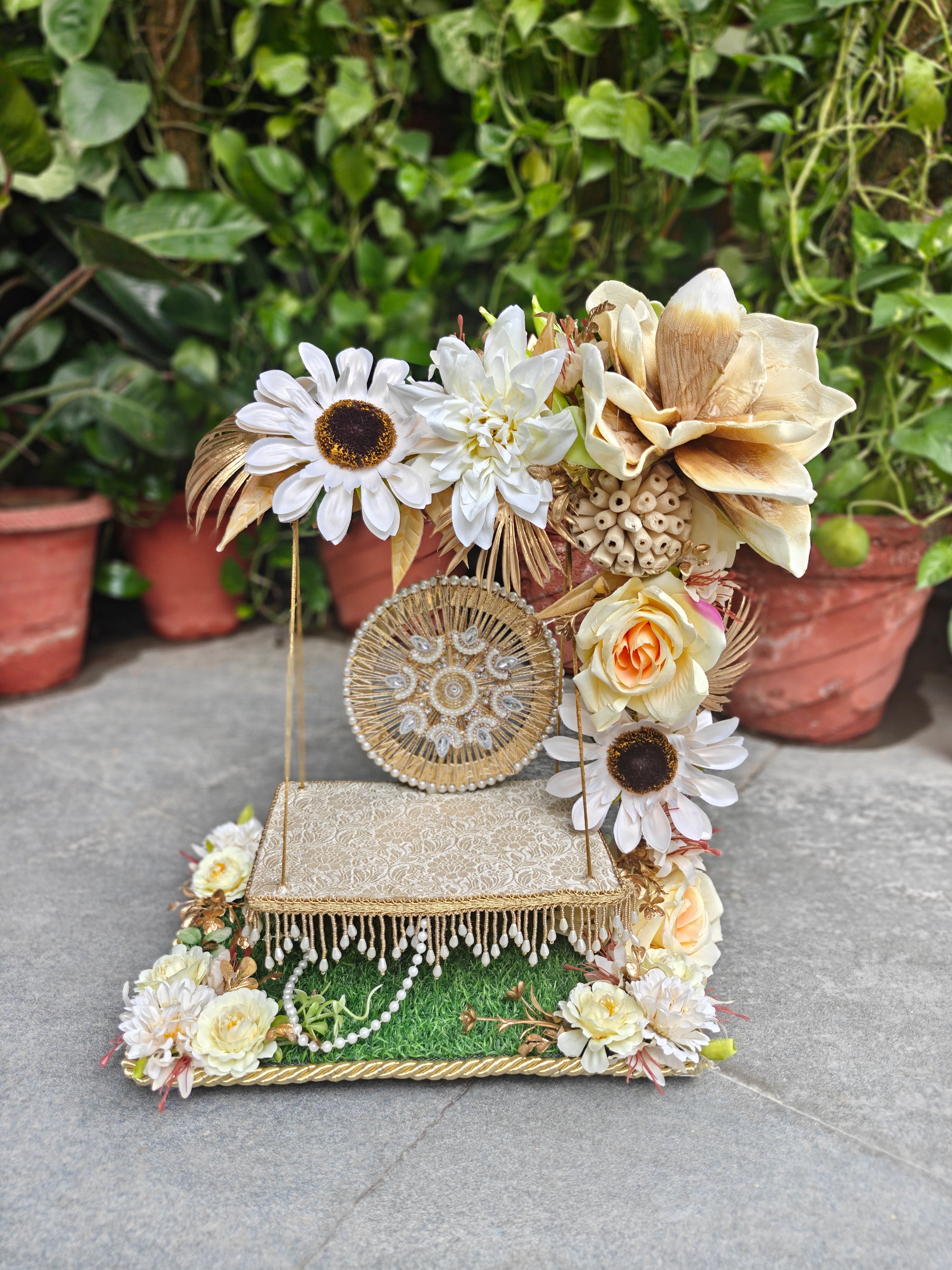 A decorative floral display stand with faux flowers, a small woven basket, faux pearls, and a lace-trimmed platform, photographed outdoors on a stone surface with potted plants in the background.