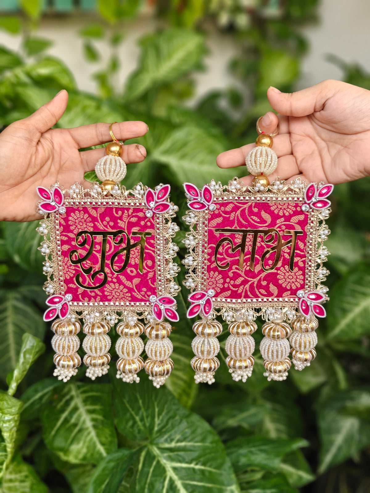Pair of pink brocade Shubh Labh hangings with gold acrylic lettering, gota patti border, pink lotus corners and pearl latkans held in hand