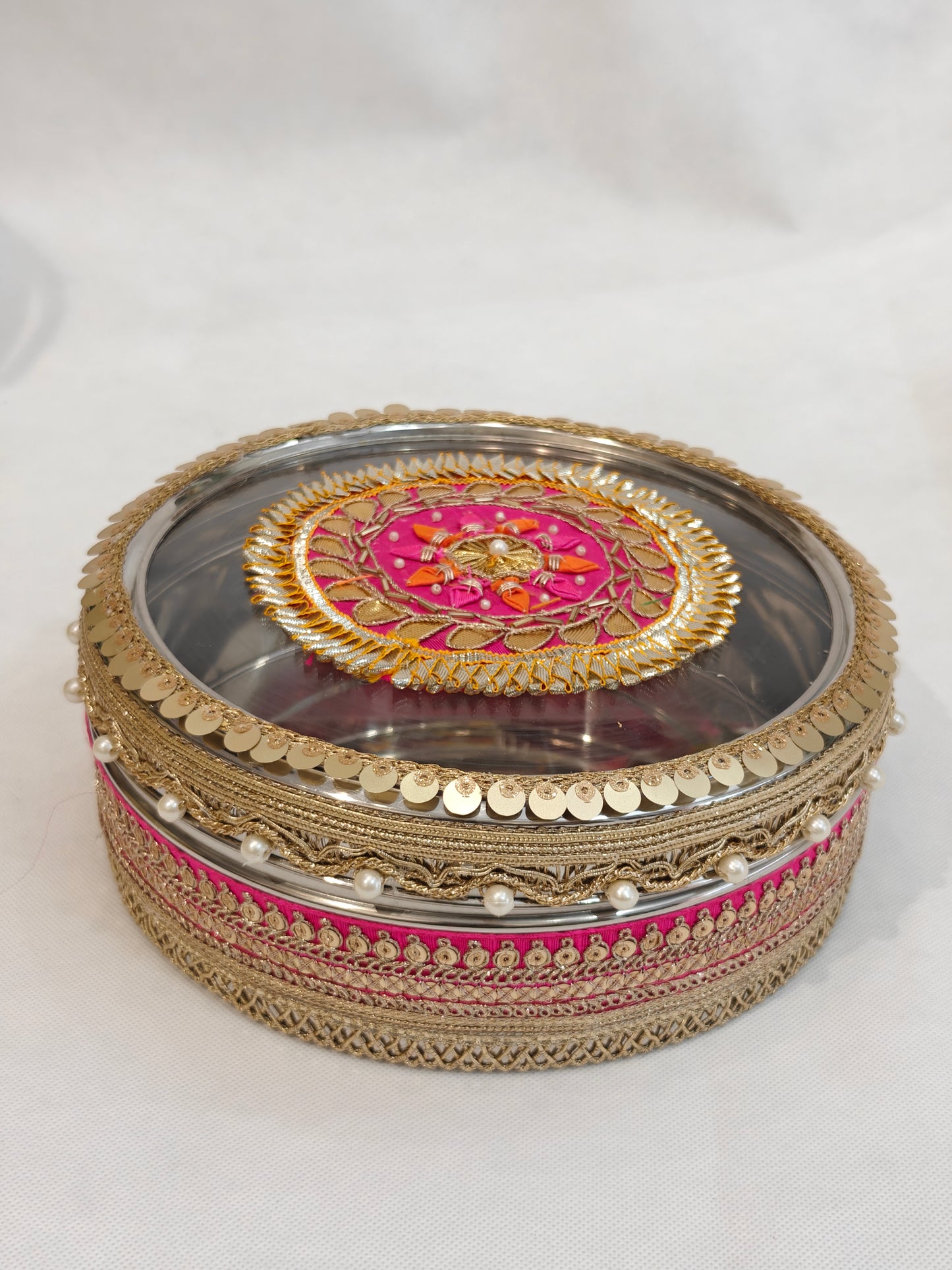 Decorated stainless steel round storage box with transparent acrylic lid featuring pink and gold zardozi medallion, pearl drops, gold coin sequin border and zari lace trim for Indian wedding trousseau