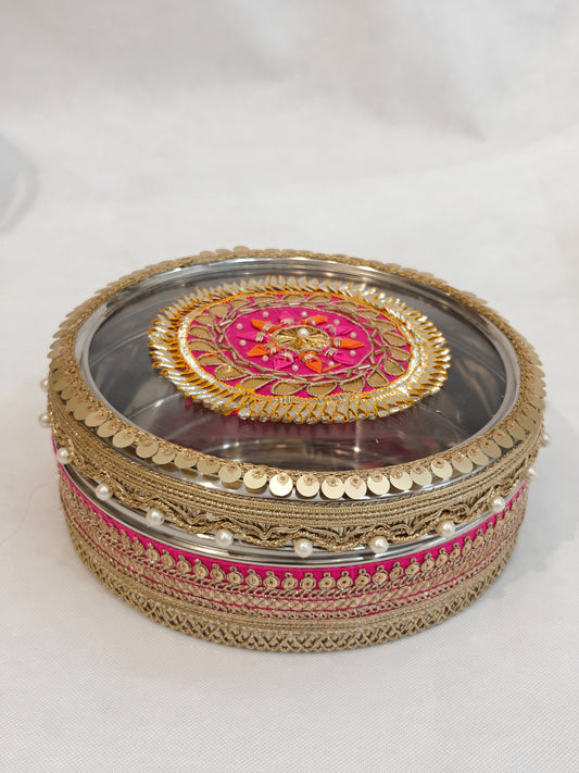 Decorated stainless steel round storage box with transparent acrylic lid featuring pink and gold zardozi medallion, pearl drops, gold coin sequin border and zari lace trim for Indian wedding trousseau