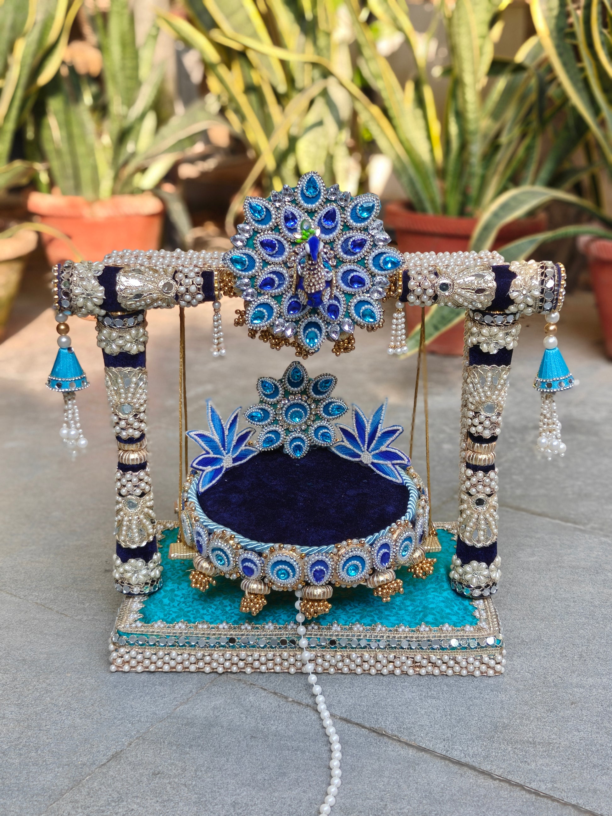 Front view of Royal Collection 2-in-1 handcrafted Ladoo Gopal peacock jhula in royal navy blue and teal velvet – 3D enamel peacock torana crown with sapphire and aqua teardrop stones, navy and silver zardozi pillar frame with pearl clusters, sky blue ghungroo corner bells, detachable navy patra with peacock lotus backrest and gold ghungroo legs, teal MDF base with double pearl border – The Wedding Station