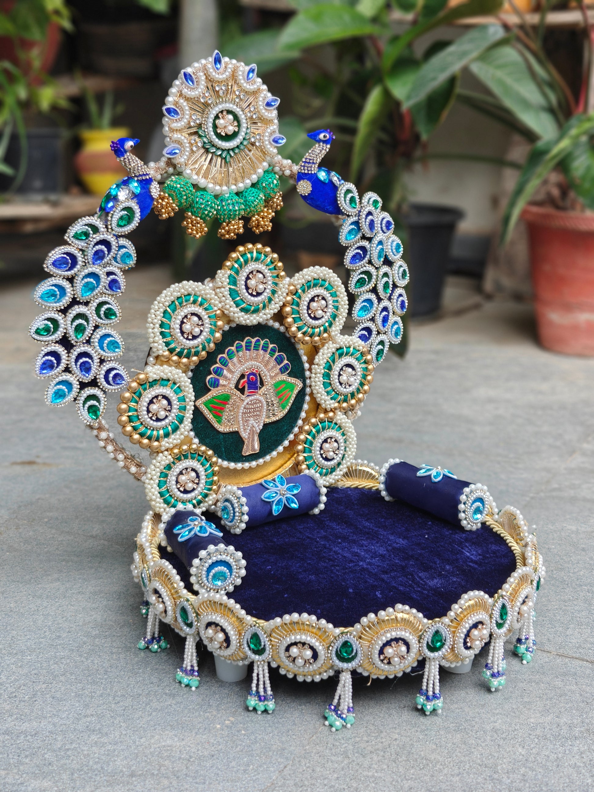 An ornate decorative throne chair featuring a deep navy blue velvet seat cushion, heavily embellished with gold, white pearls, and green and blue beadwork, with peacock feather motifs and a central peacock emblem.