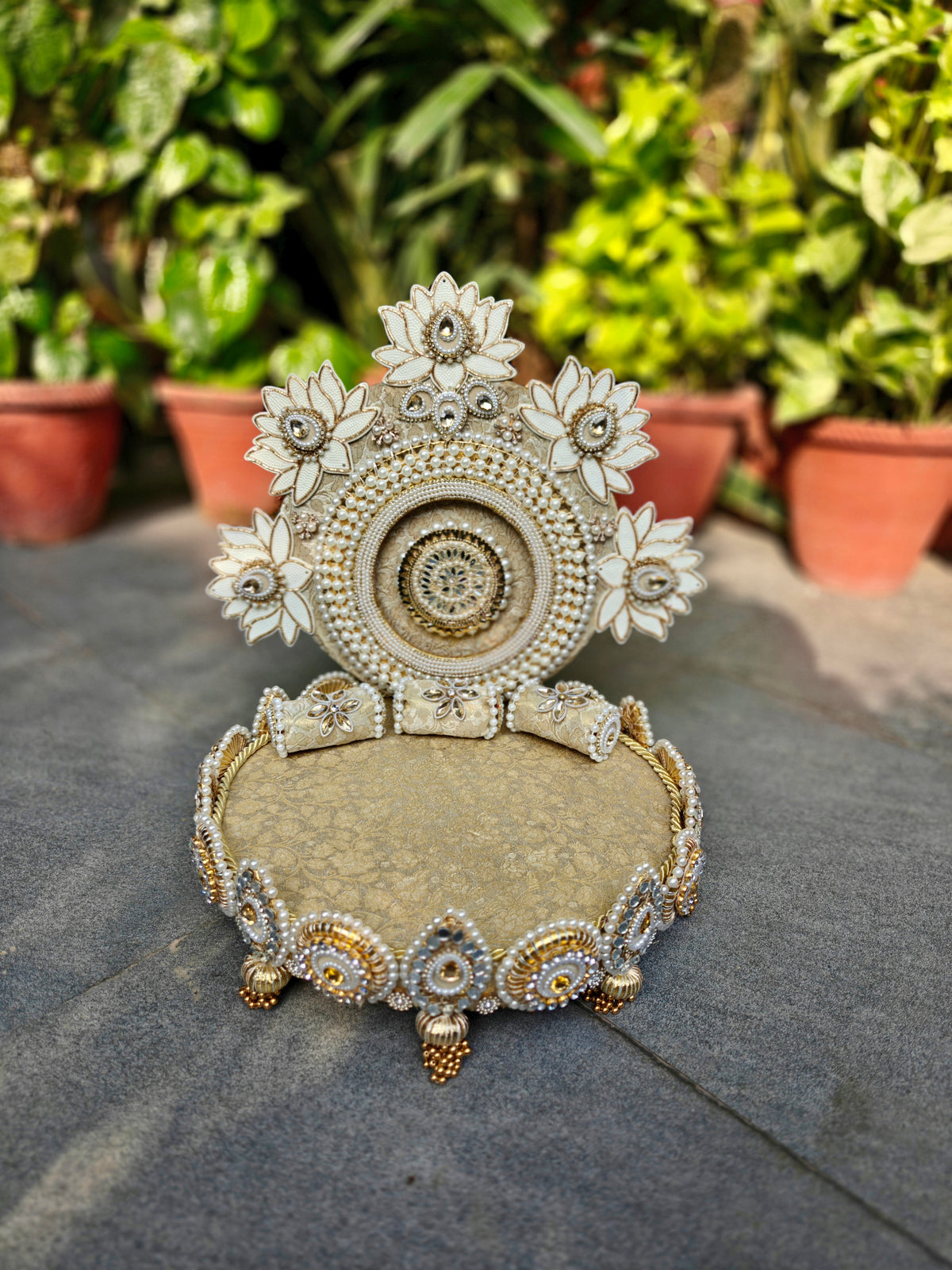 An ornate decorative jewelry display stand with a floral arch and cushion, styled outdoors on a stone surface with potted green plants in the background.