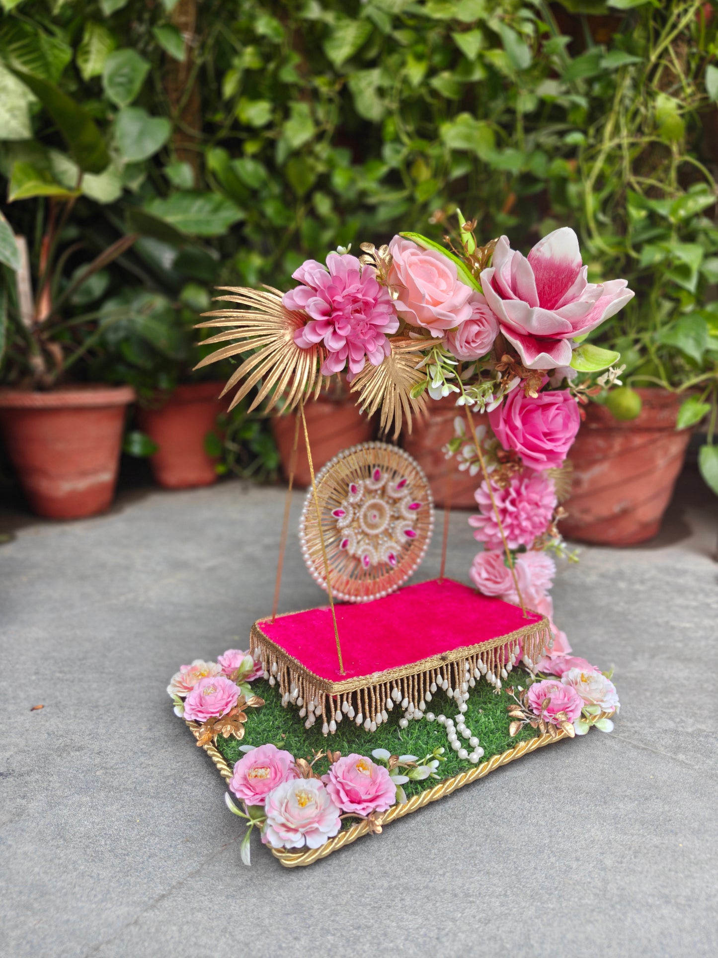 A decorative floral and ornamental display featuring pink flowers, green foliage, gold rope details, and a bright pink cushioned rectangular base, photographed outdoors on a gray stone surface with potted plants in the background.