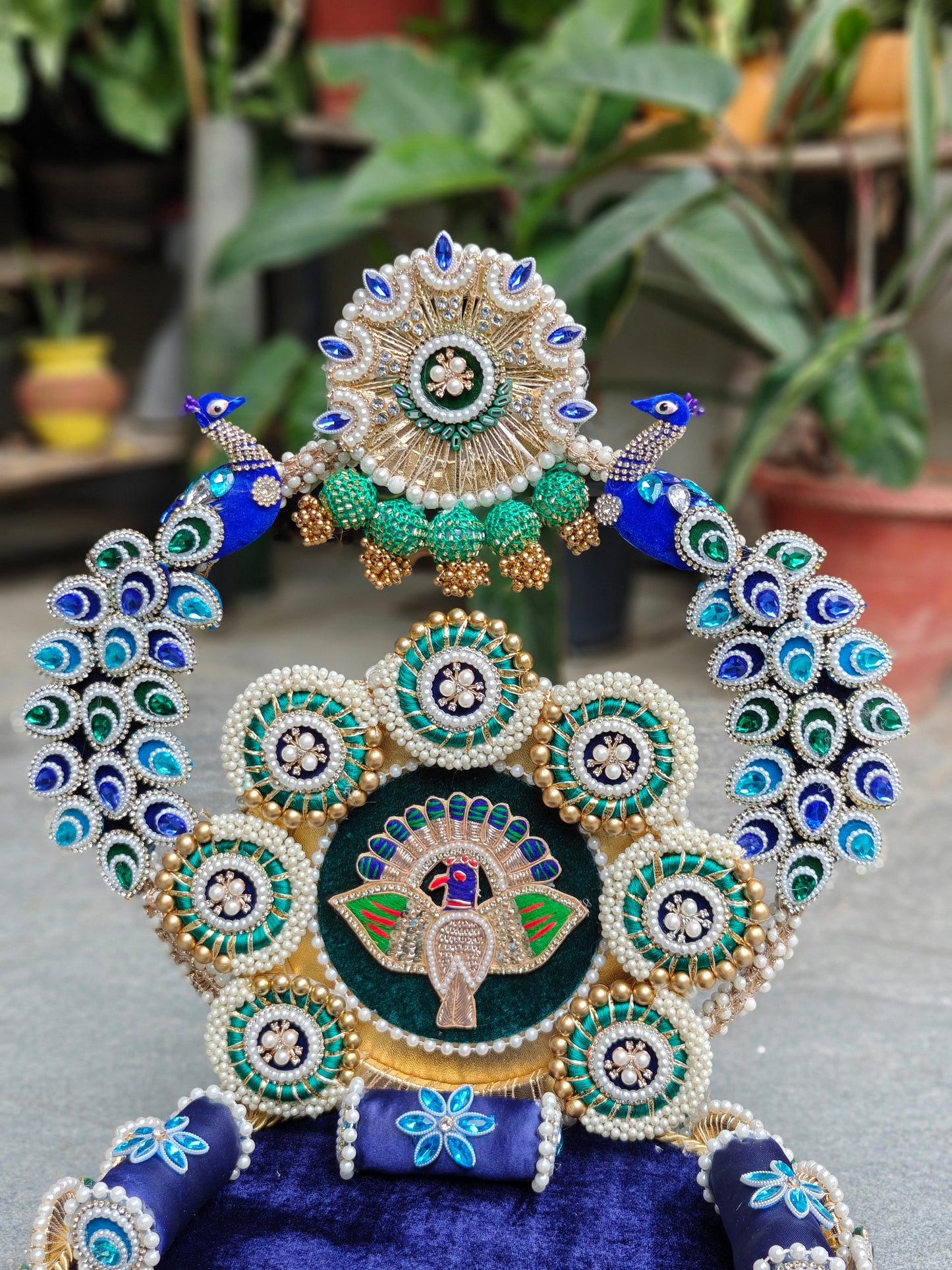 An ornate decorative peacock-themed headpiece or crown embellishment with blue fabric, white pearls, green and blue beadwork, and a central peacock motif, photographed against a blurred outdoor garden background.