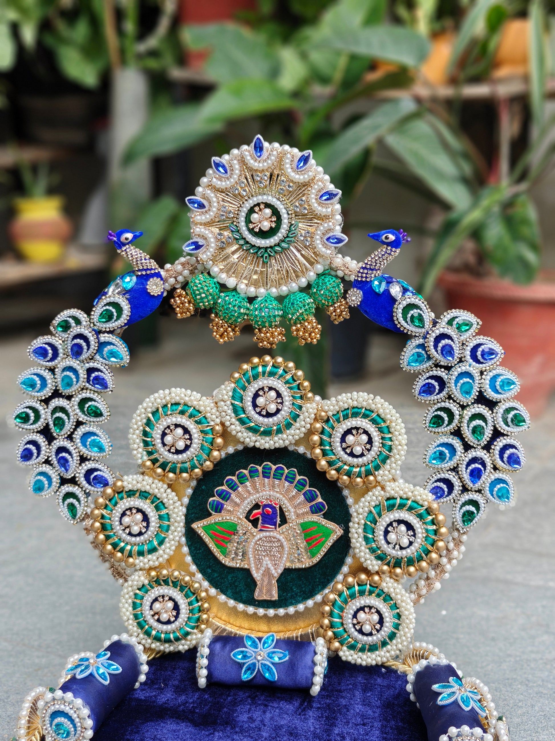 An ornate decorative peacock-themed headpiece or crown embellishment with blue fabric, white pearls, green and blue beadwork, and a central peacock motif, photographed against a blurred outdoor garden background.