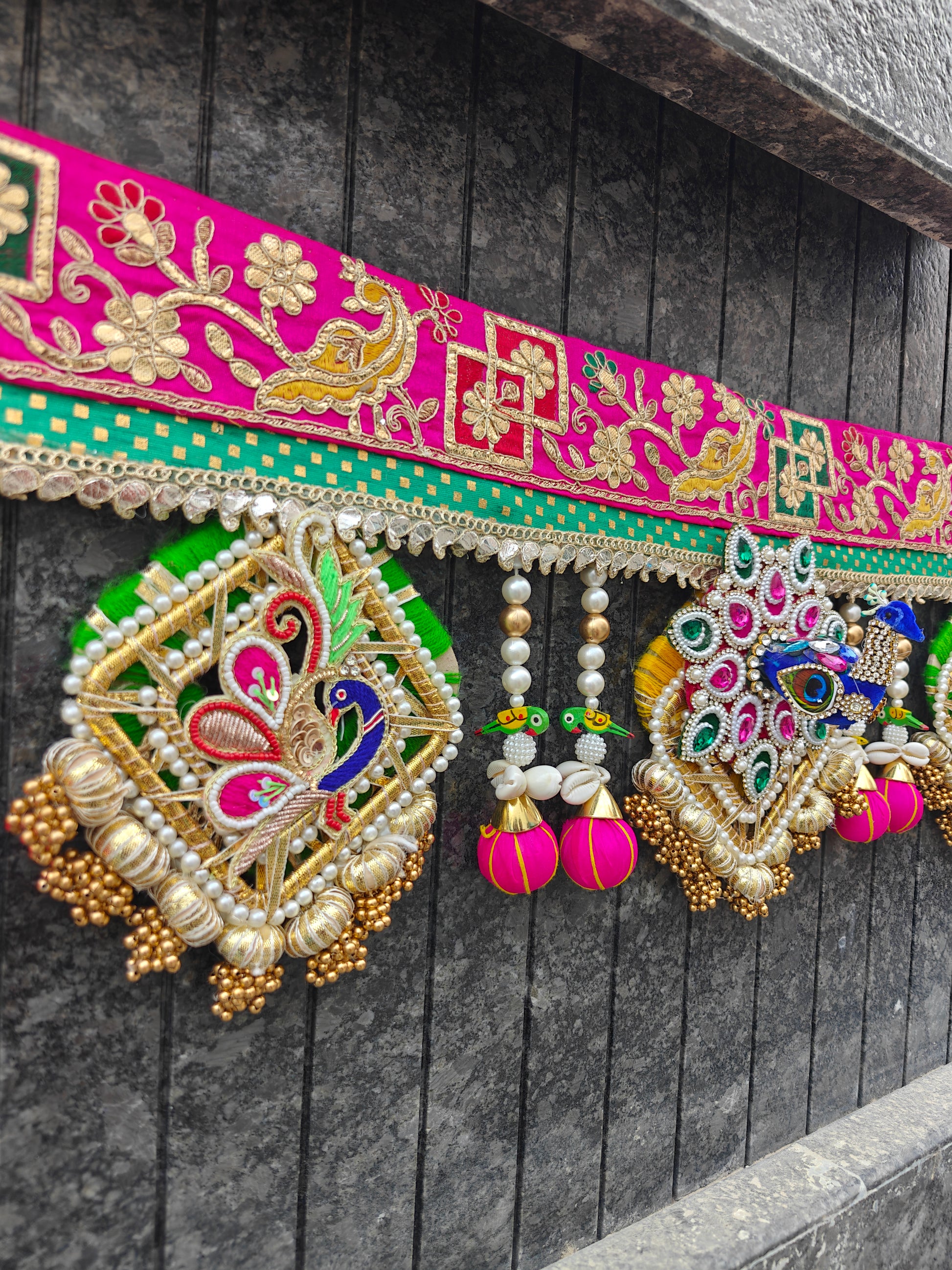 A close-up of a vibrant pink and green embroidered decorative garland with hanging ornate embellishments, beads, and tassels against a dark gray paneled wall.