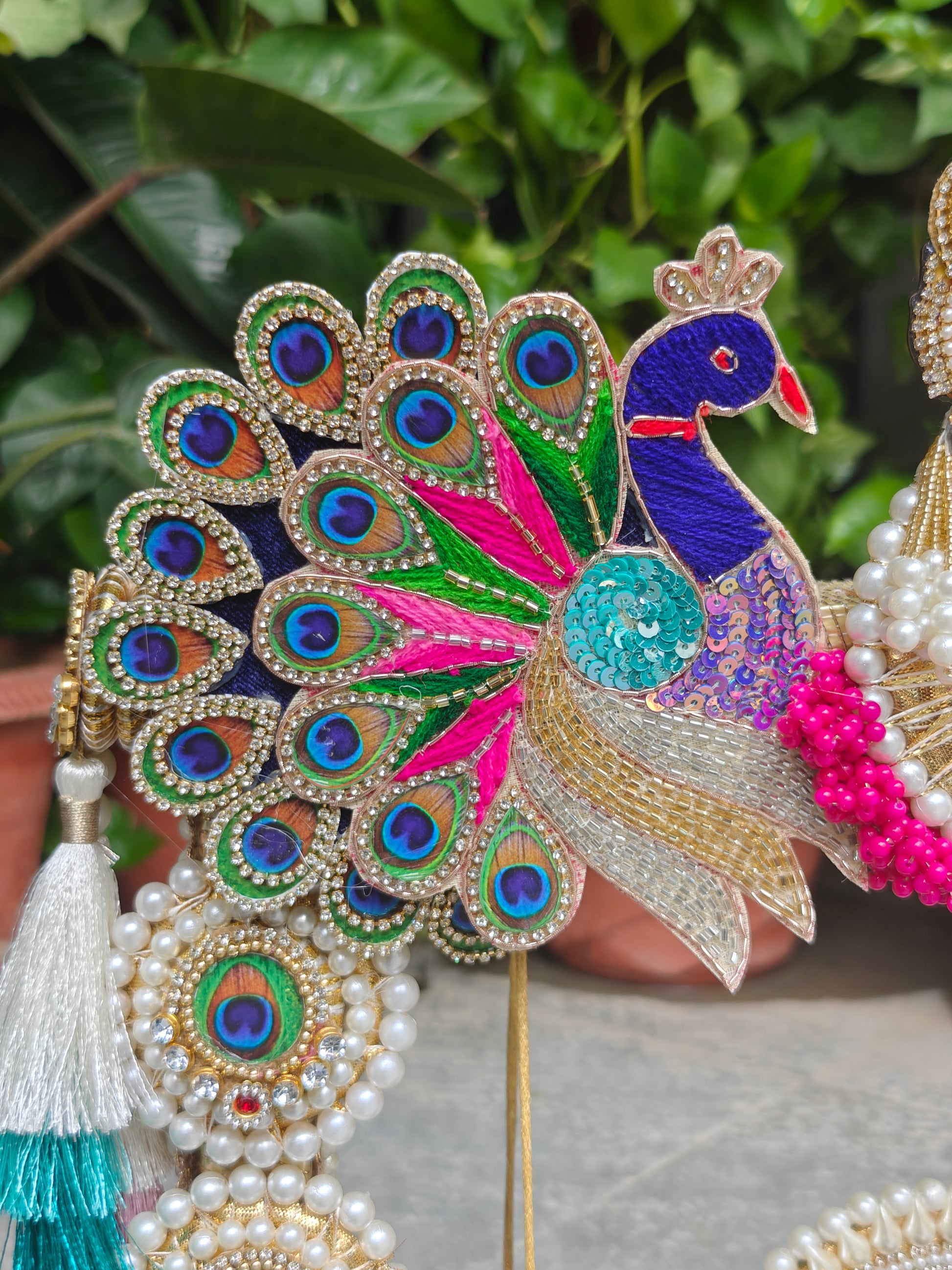 A close-up of an ornate peacock figurine or decorative piece with a fanned tail of embroidered peacock-eye feathers, a blue peacock body, bright pink and green accents, and dangling pearl strands and tassels, set against a blurred green foliage background.