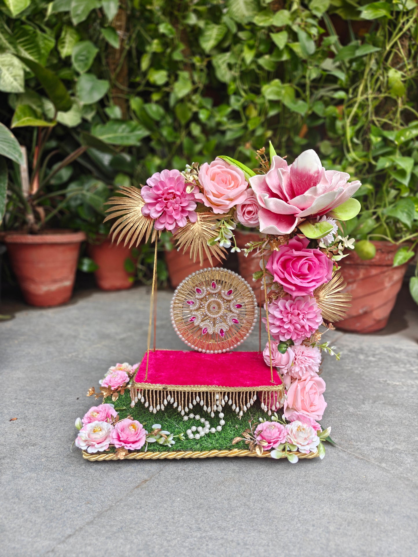 A decorative floral and textile display stand with pink flowers, a bright pink fabric-covered platform, gold trim, and a round embroidered panel, photographed outdoors against a background of potted green plants.