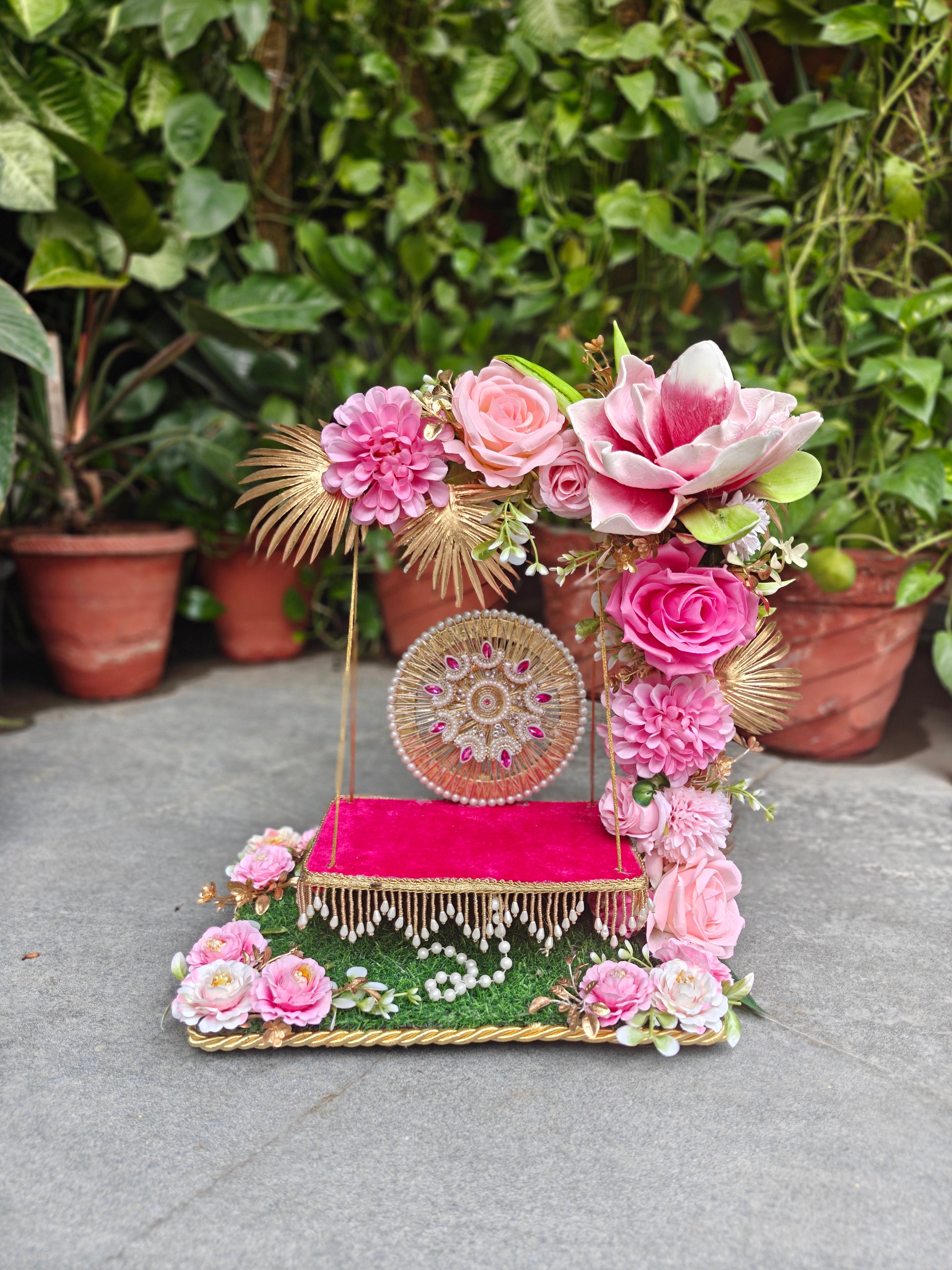 A decorative floral and textile display stand with pink flowers, a bright pink fabric-covered platform, gold trim, and a round embroidered panel, photographed outdoors against a background of potted green plants.