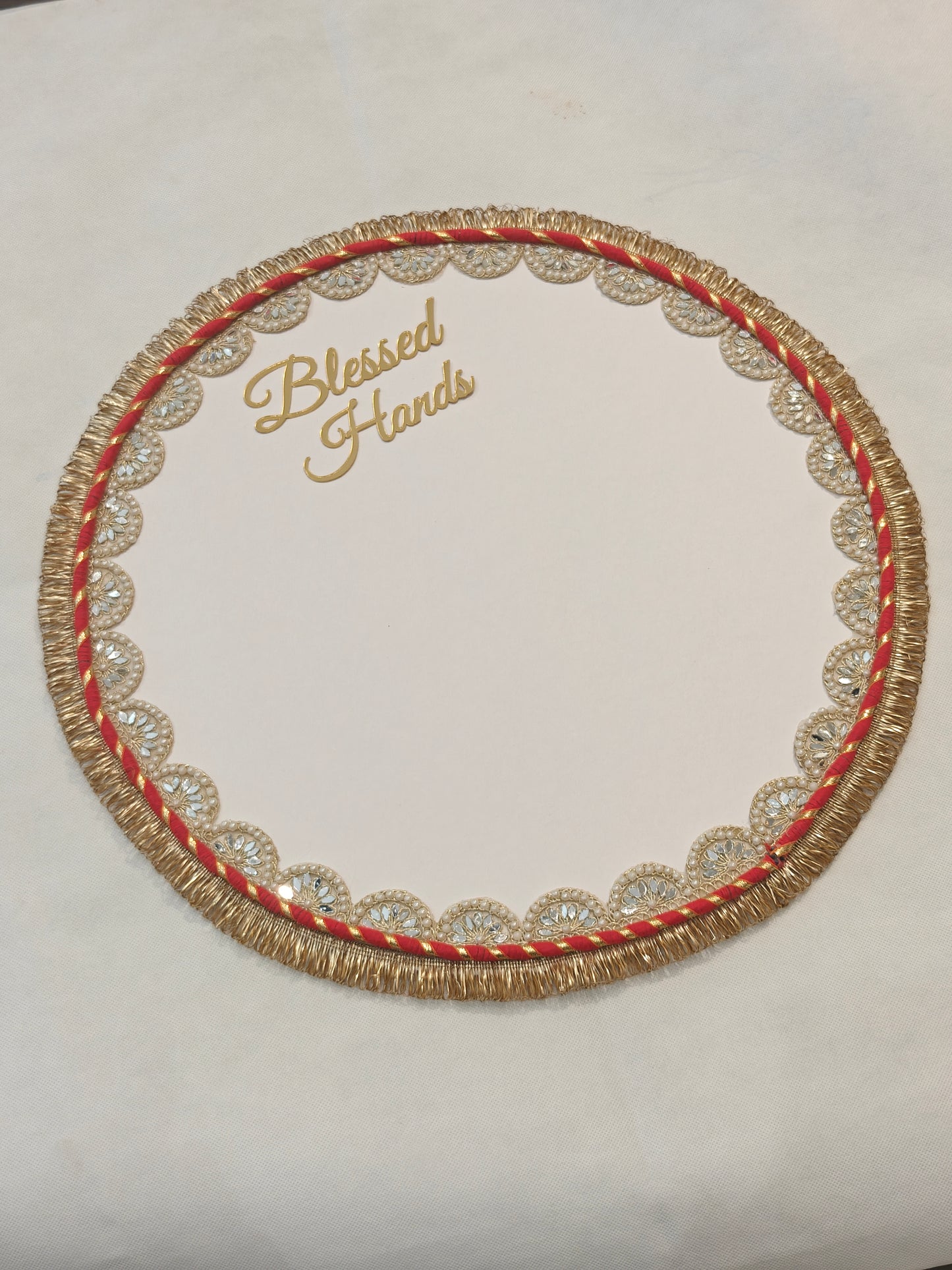 Handcrafted 16-inch round white handprint mat with gold acrylic "Blessed Hands" script, gold zari fringe border, red and gold twisted rope band and pearl mirror scallop lace trim on MDF base — Griha Pravesh and shop opening ceremony mat by The Wedding Station Meerut