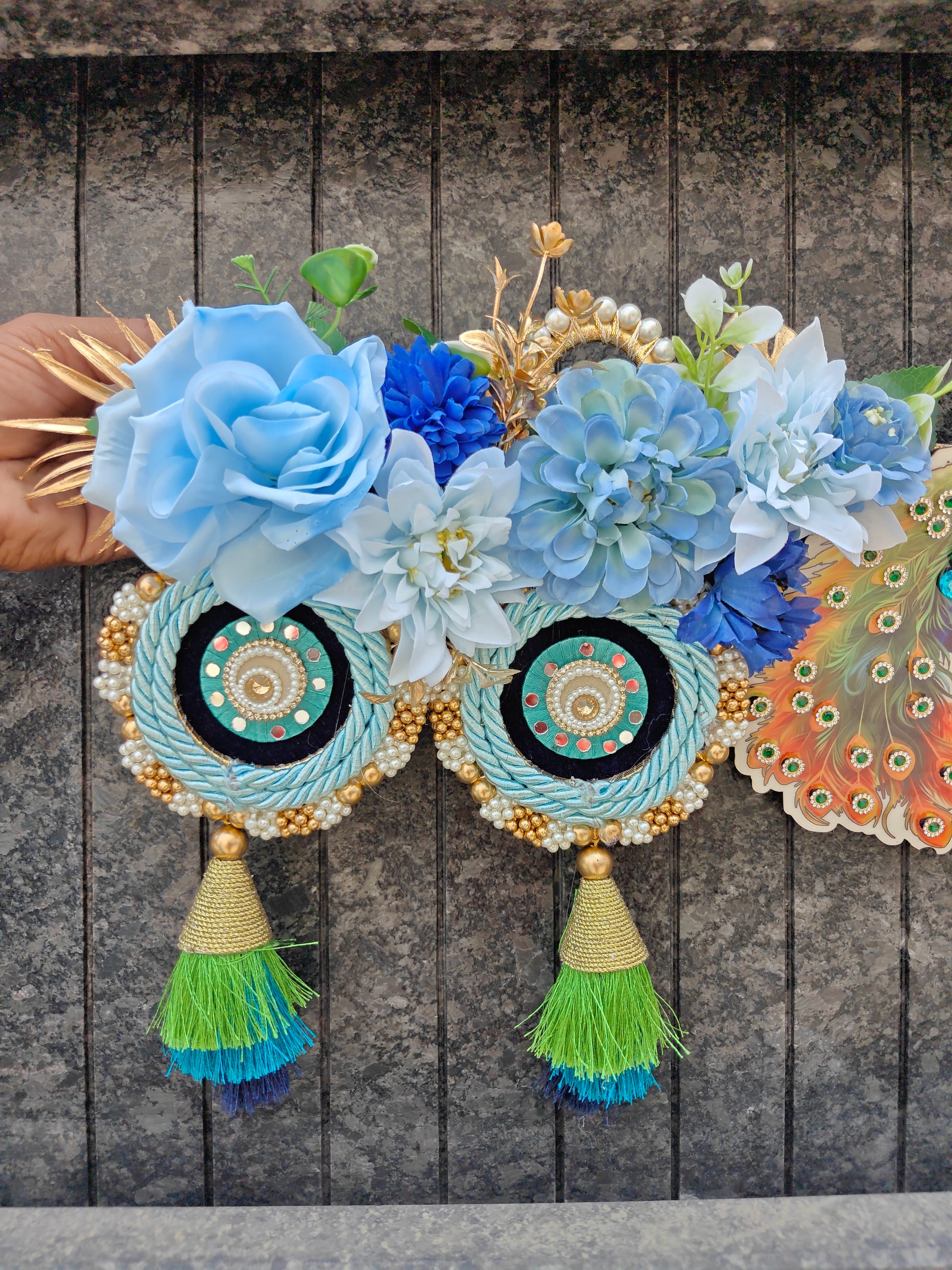 A decorative garland with large blue and white fabric flowers, two circular woven medallions with turquoise and gold patterns, and two tassels in green and blue, held against a dark wood plank background.