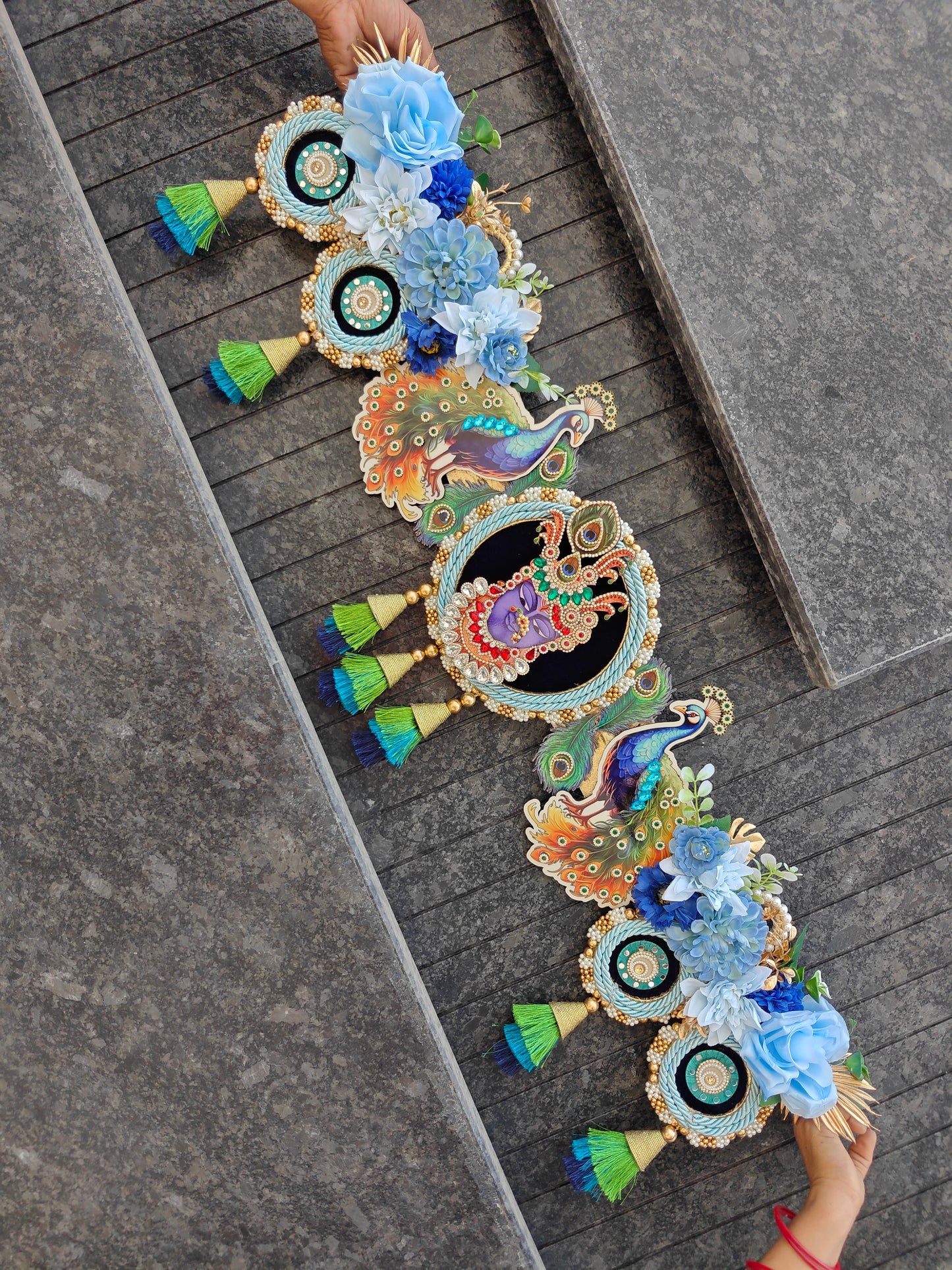 A decorative garland laid diagonally across gray stone steps, featuring peacock motifs, floral clusters in blue and white, and small green-and-blue tassels. Two hands are visible holding the ends of the garland.