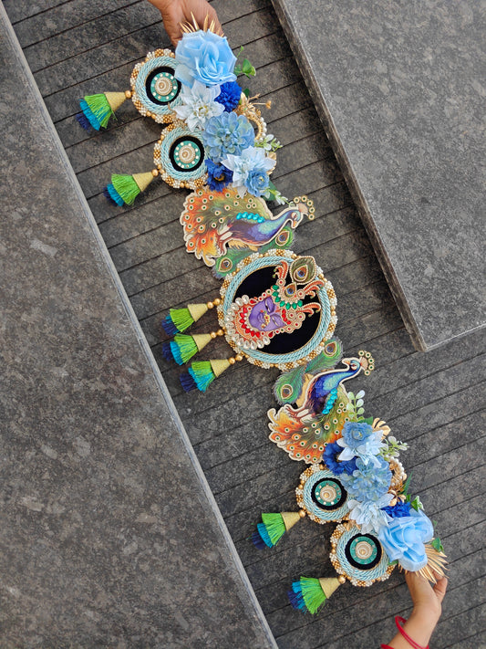 A decorative garland laid diagonally across gray stone steps, featuring peacock motifs, floral clusters in blue and white, and small green-and-blue tassels. Two hands are visible holding the ends of the garland.