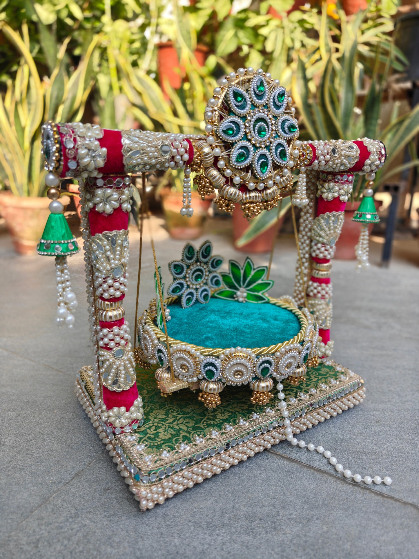 Side angle view of Royal Collection 2-in-1 handcrafted Ladoo Gopal jhula in emerald green and crimson velvet – brocade MDF base with mirror border, crimson pillar frame with pearl clusters, teal velvet detachable patra with lotus backrest and emerald torana crown – The Wedding Station