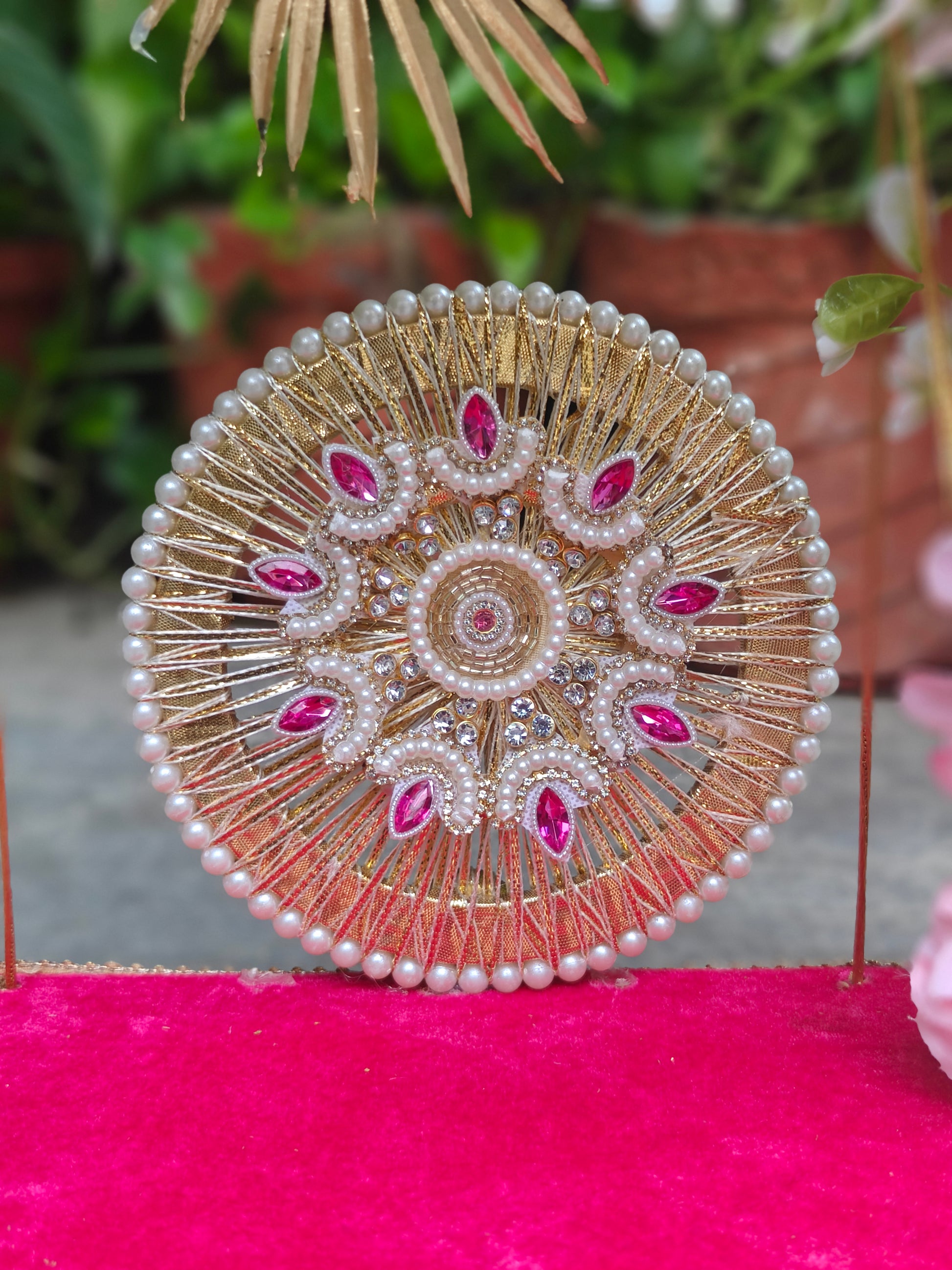 A circular gold-toned ornamental jewelry stand or decorative display piece with rows of white faux pearls and pink gemstone accents, photographed on a bright pink surface with blurred greenery in the background.