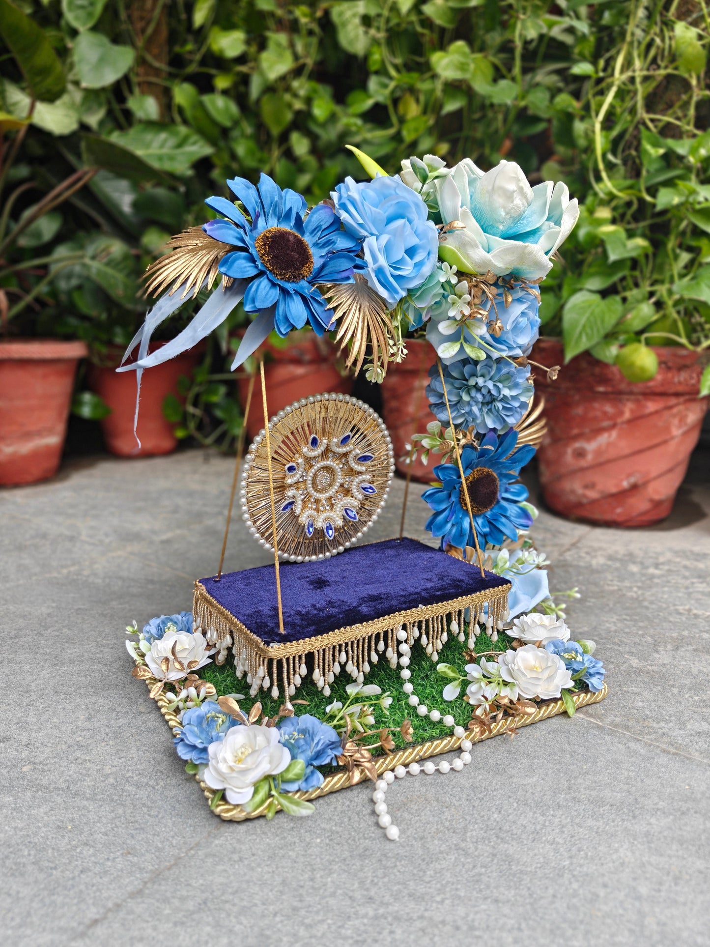 A decorative floral display stand with a purple velvet base, gold trim, blue and white artificial flowers, and a round gold medallion, photographed outdoors on a stone surface with potted plants in the background.