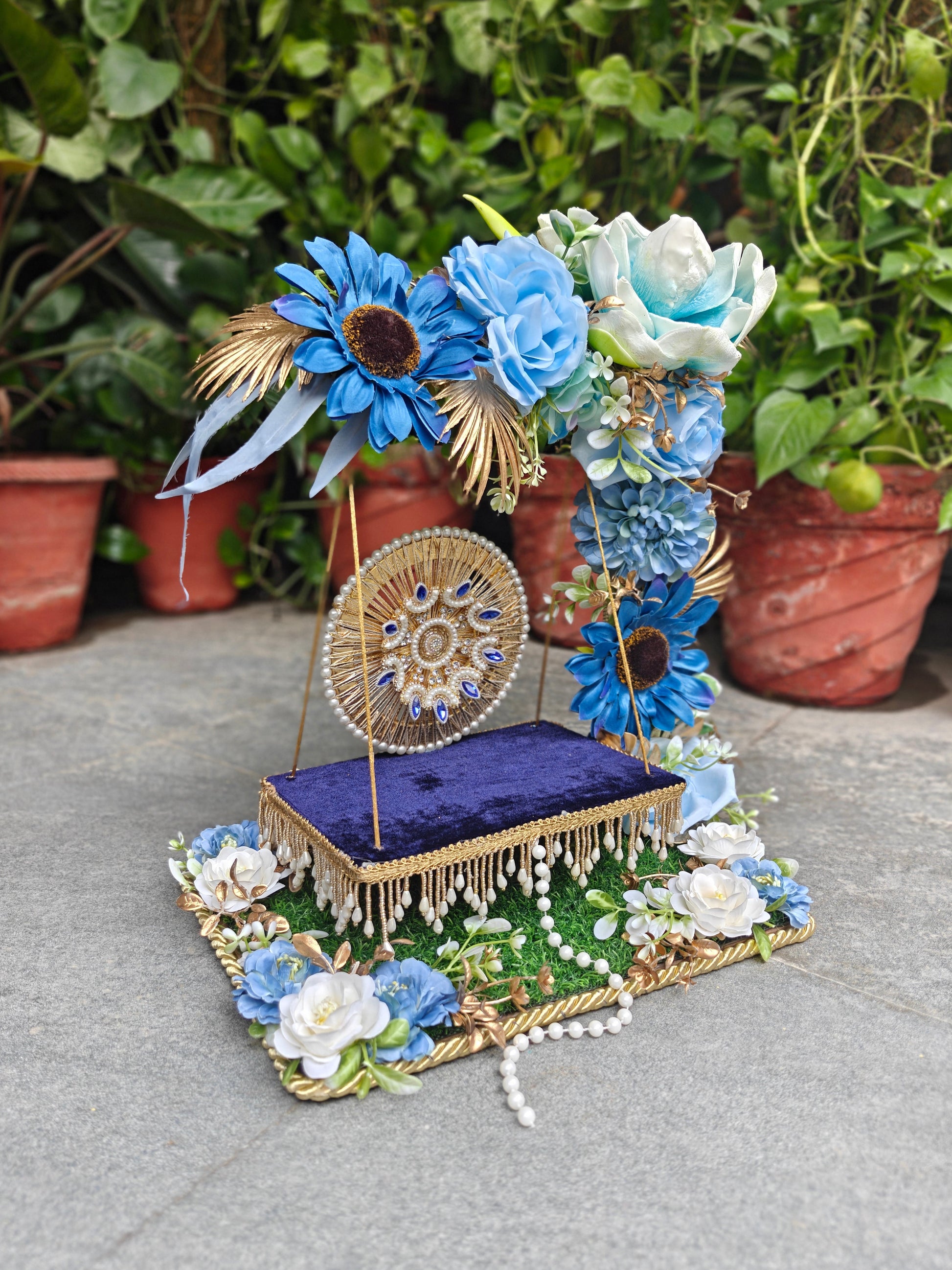 A decorative floral display stand with a purple velvet base, gold trim, blue and white artificial flowers, and a round gold medallion, photographed outdoors on a stone surface with potted plants in the background.