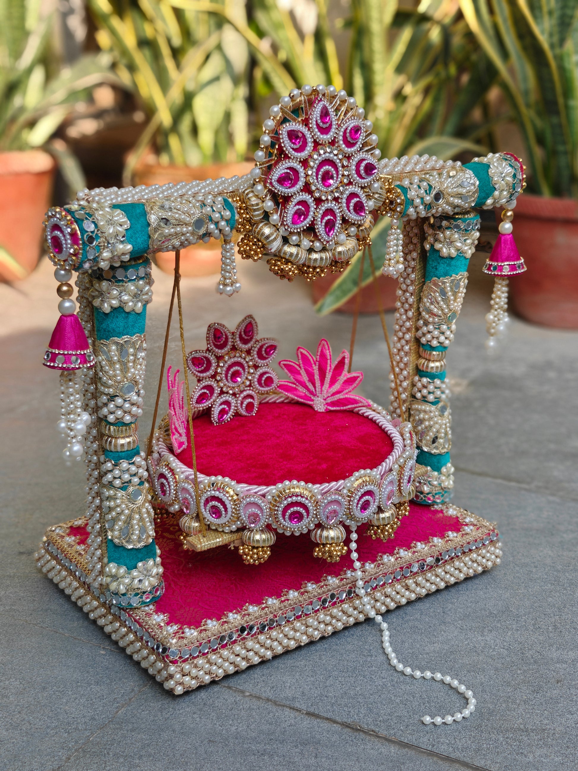 Royal Collection 2-in-1 handcrafted Ladoo Gopal jhula and singhasan in rani pink and teal velvet – decorative torana crown with fuchsia teardrop stones, teal pillar frame with mirror work and pearl clusters, detachable lotus patra singhasan with gold ghungroo fringe, MDF base with mirror border and pearl edging – The Wedding Station